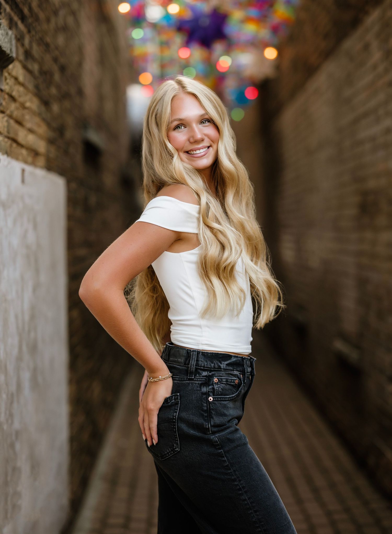 Person with long blonde wavy hair poses in a white top and dark jeans against brick walls with colorful bokeh lights.