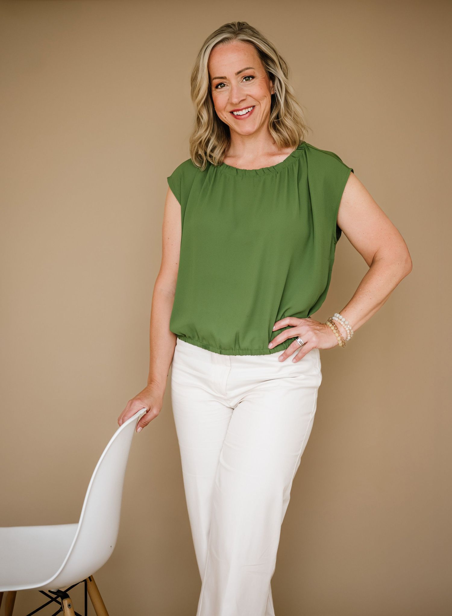 Professional portrait shot against beige backdrop showing a green sleeveless blouse paired with white pants and modern styling.