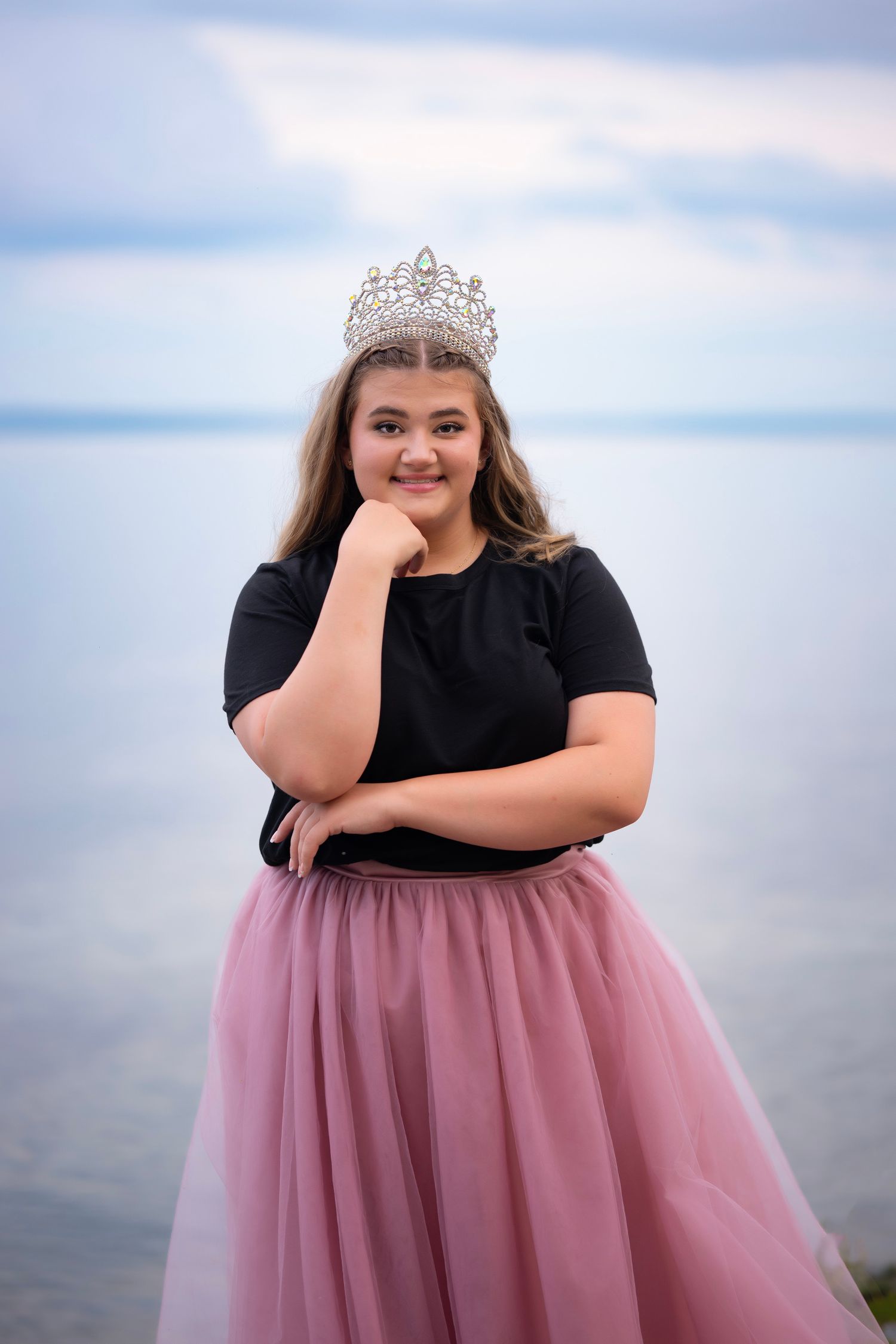 Young woman wearing a rhinestone crown and pink tulle skirt poses confidently by the ocean.