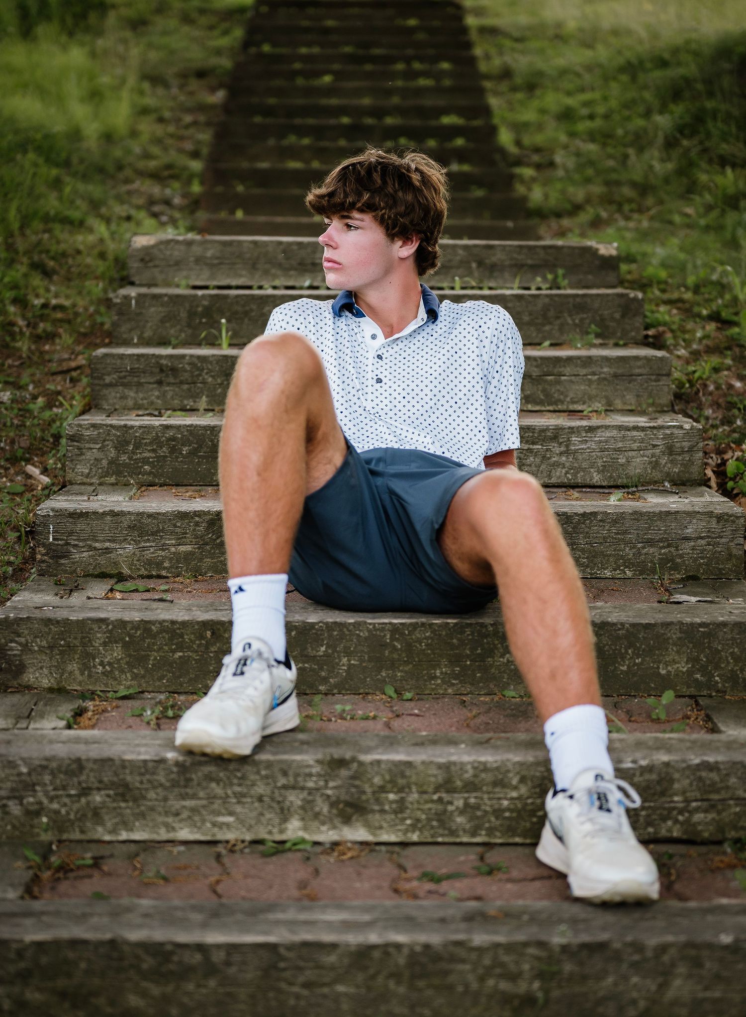 A person in a polka dot shirt and shorts sits casually on concrete steps surrounded by greenery.