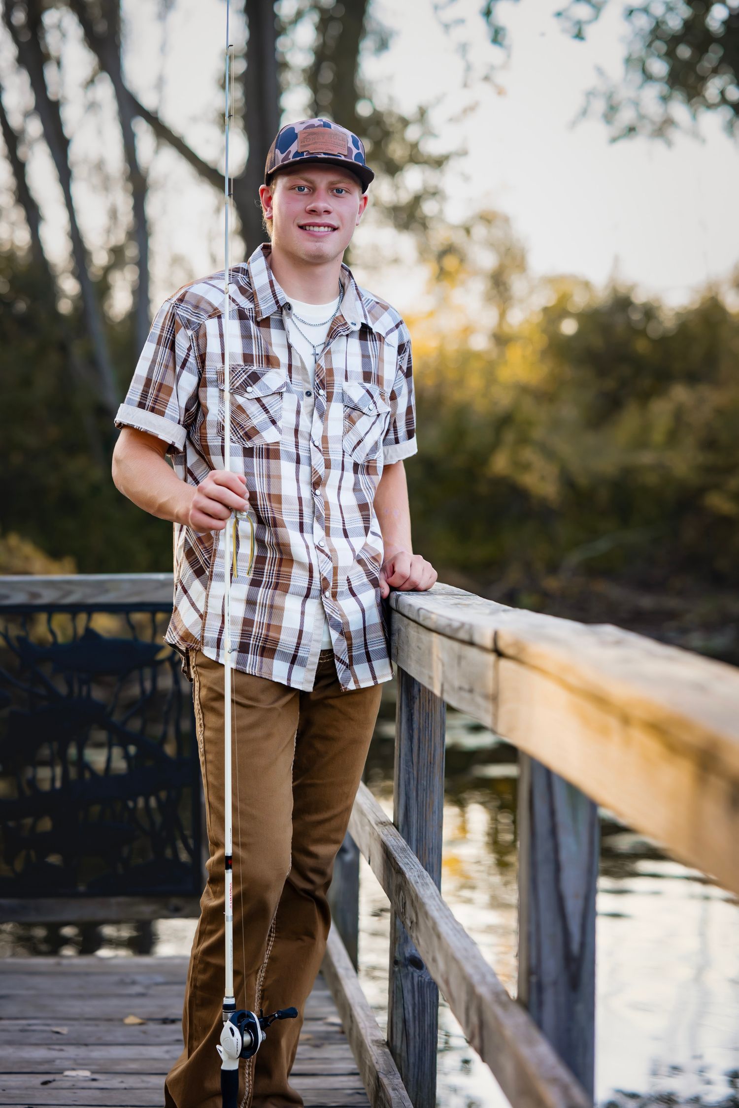 Person in plaid shirt and baseball cap leans on wooden dock railing during golden sunset by lake.