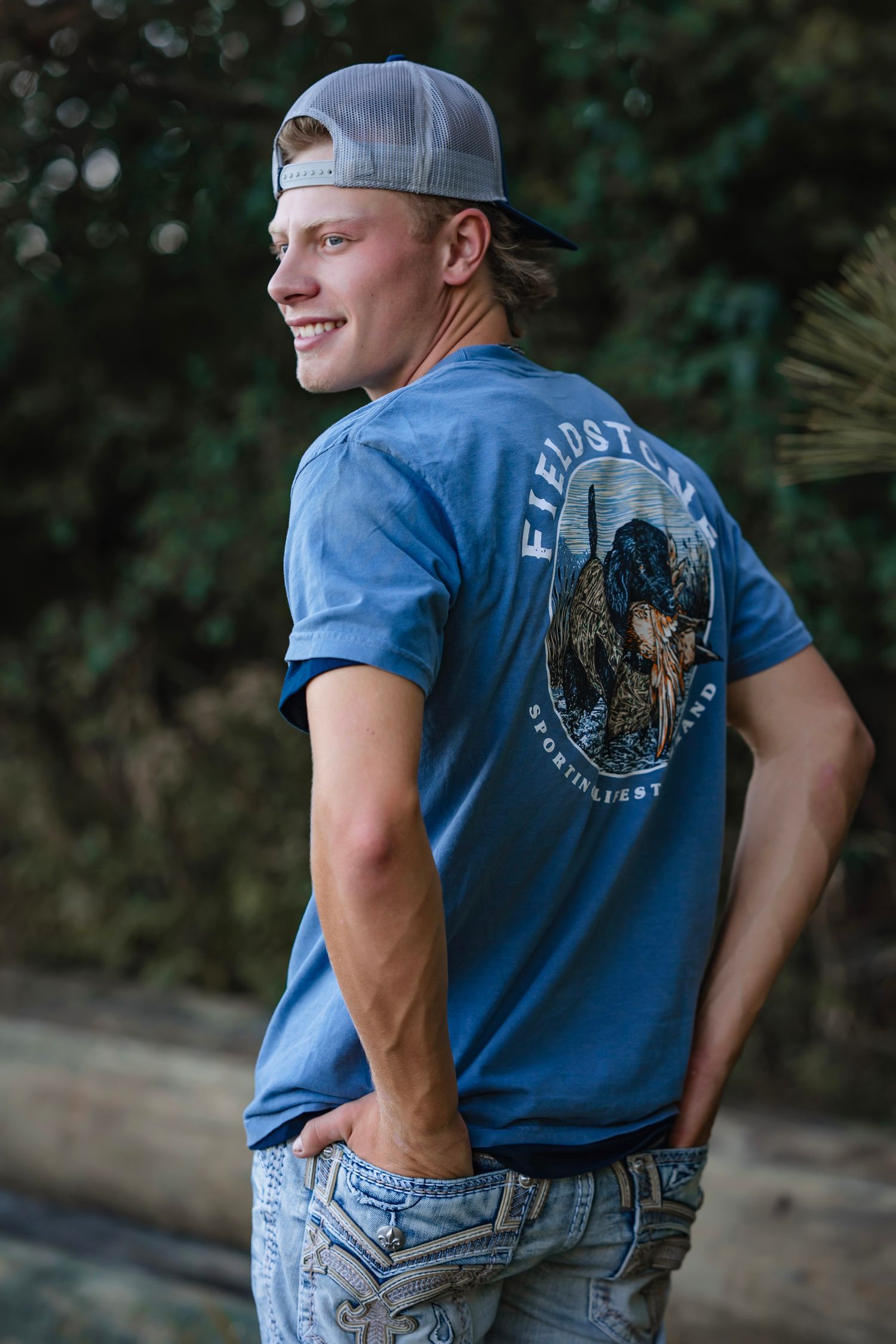 Person wearing blue t-shirt and jeans poses against blurred outdoor background in casual fashion photo series.