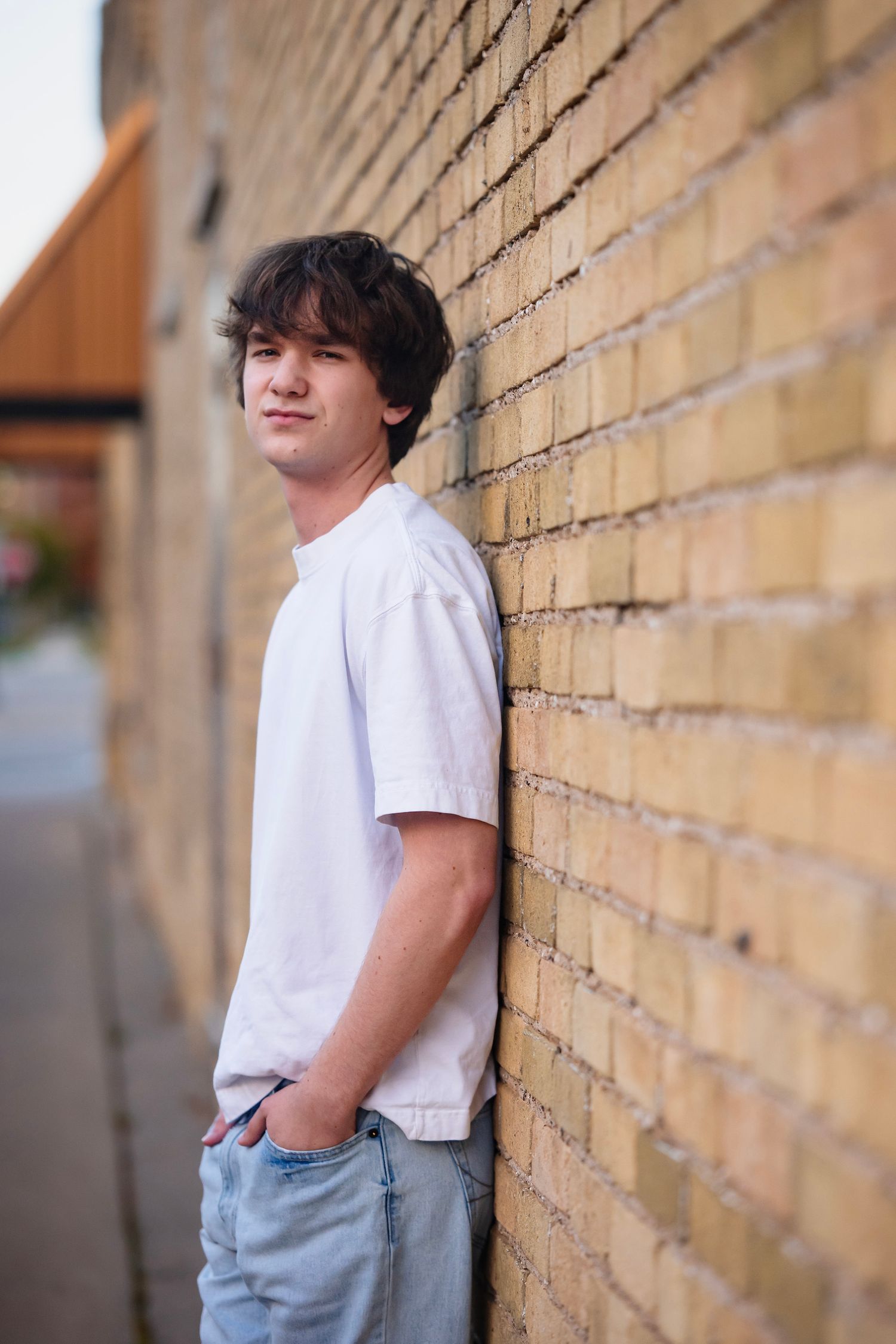 A young person in a white t-shirt and jeans leans casually against a yellow brick wall outdoors.