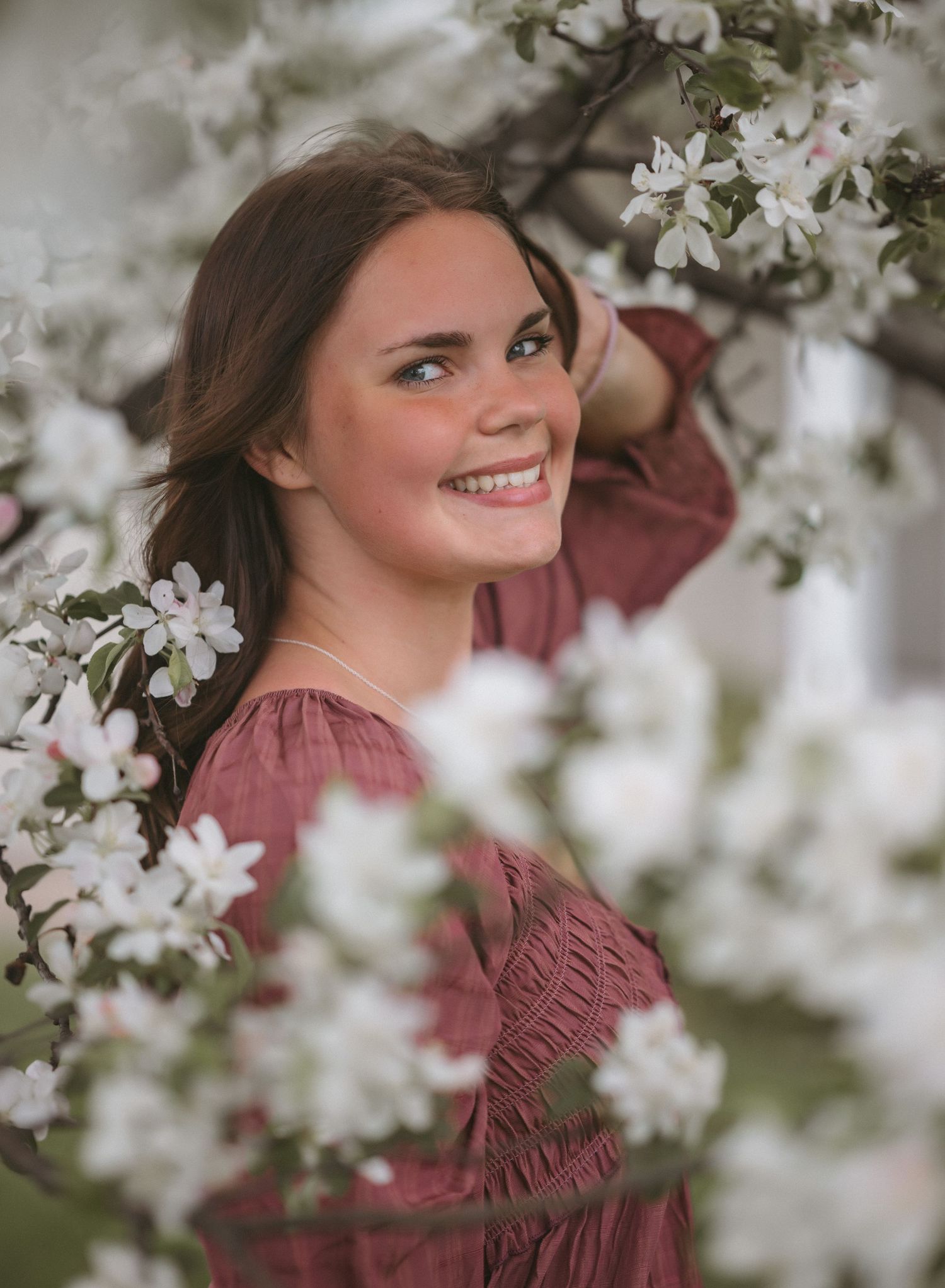 Young woman in pink dress smiling among white spring blossoms on flowering tree branches outdoors.