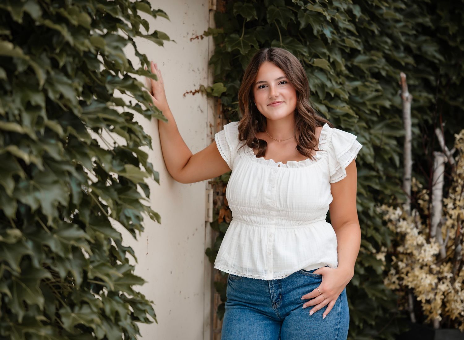 Young woman in white ruffled top and jeans poses against ivy-covered wall outdoors in natural light.
