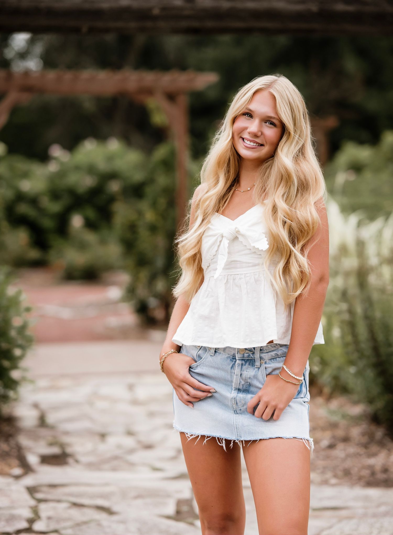 Smiling blonde teen in white bow top and denim mini skirt poses on stone path in lush garden setting.