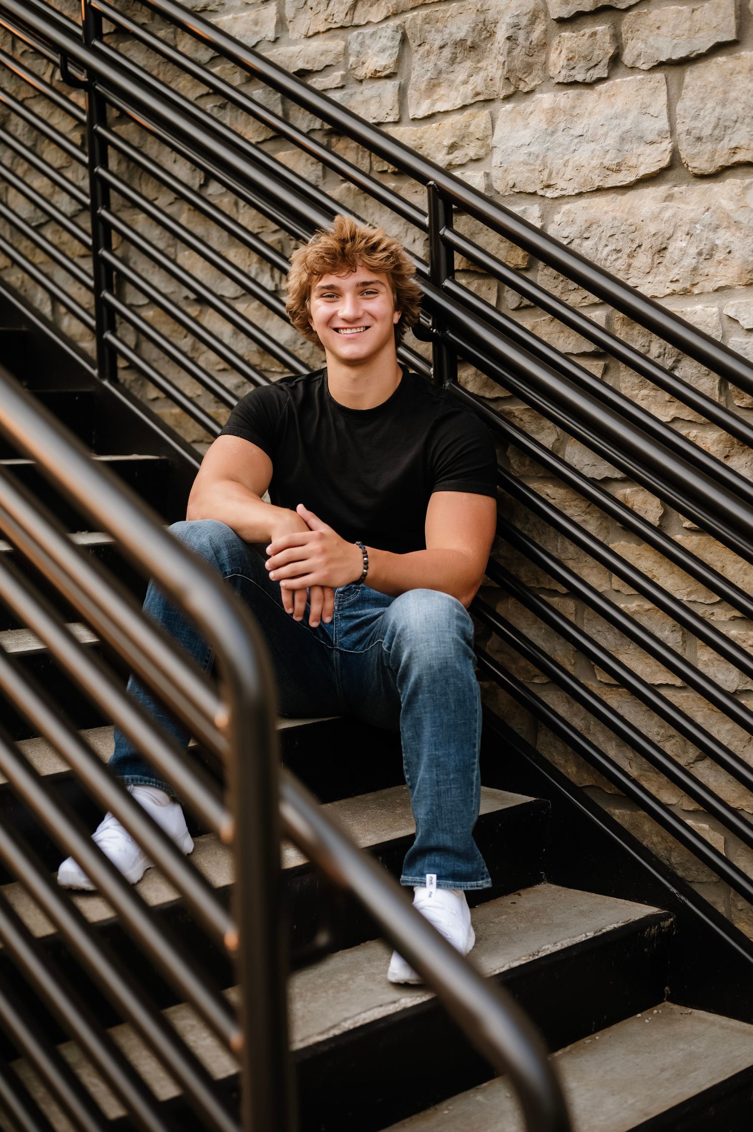 Smiling teenage boy with curly blonde hair sits on outdoor stairs wearing black t-shirt and jeans.