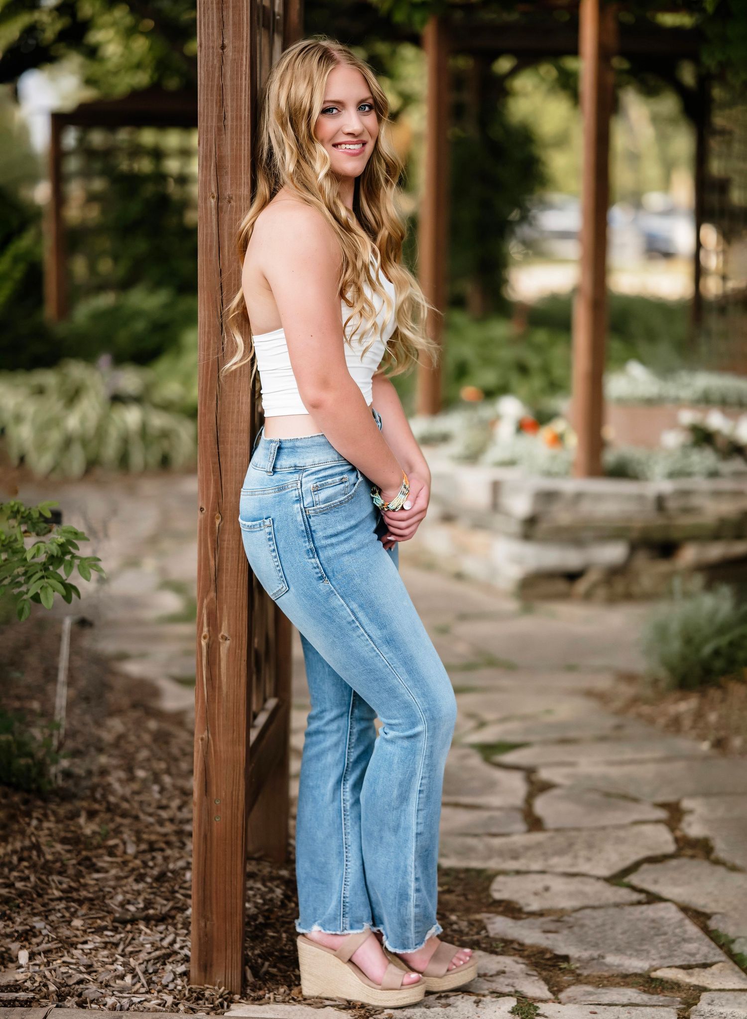 Woman in white top and light jeans sits on stone pathway surrounded by greenery and wooden posts.