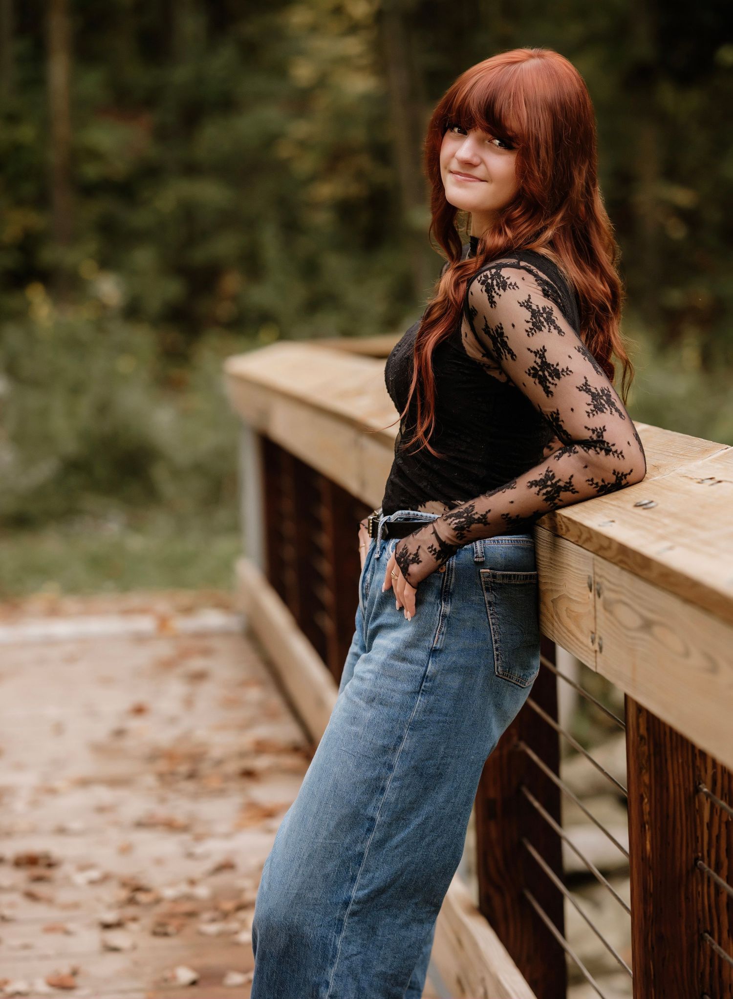 Person in black lace top and denim jeans leans against wooden railing on autumn day.