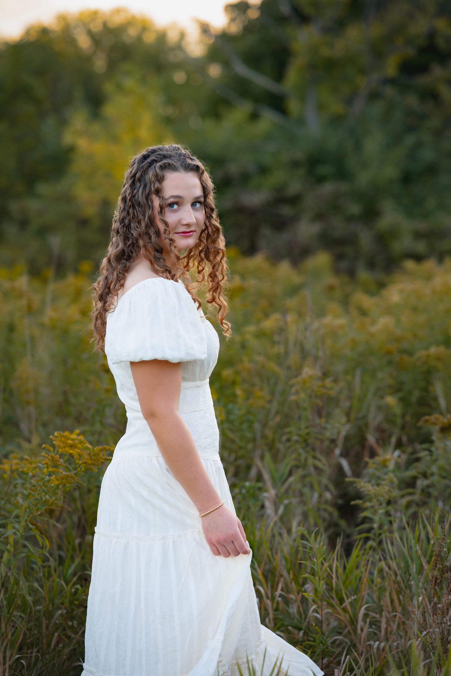 A white dress stands out against a natural backdrop of tall grass and wildflowers in golden evening light.