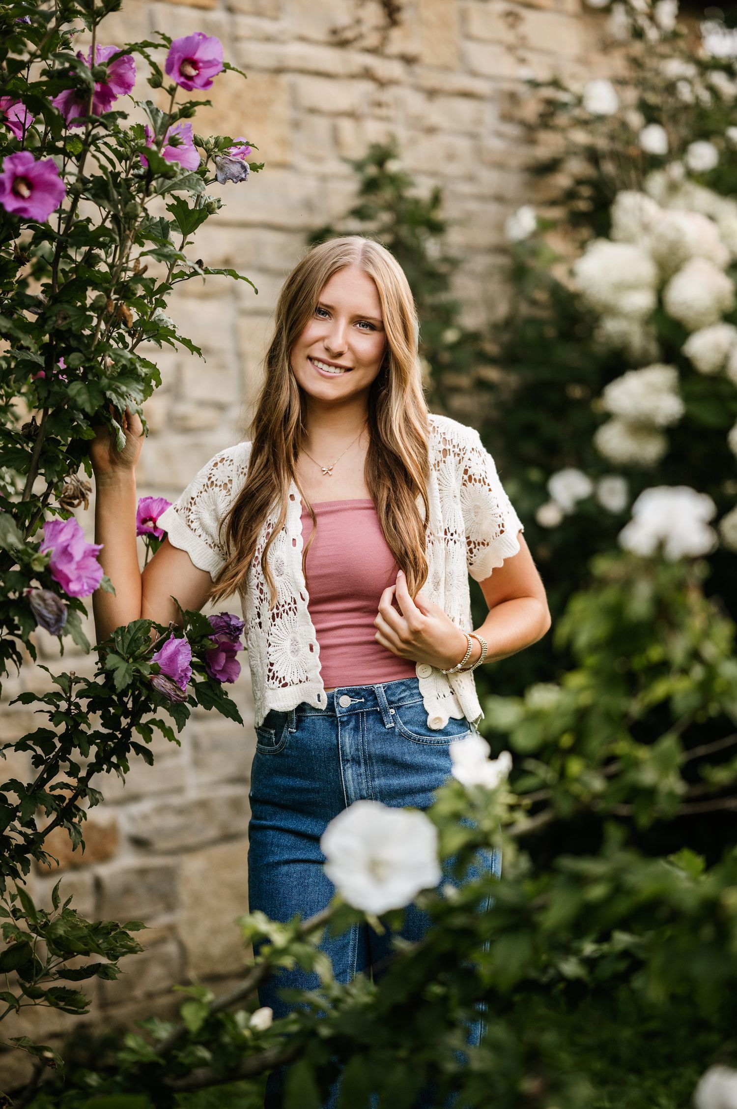Young woman in pink top and jeans smiles among blooming purple and white flowers near a stone wall.