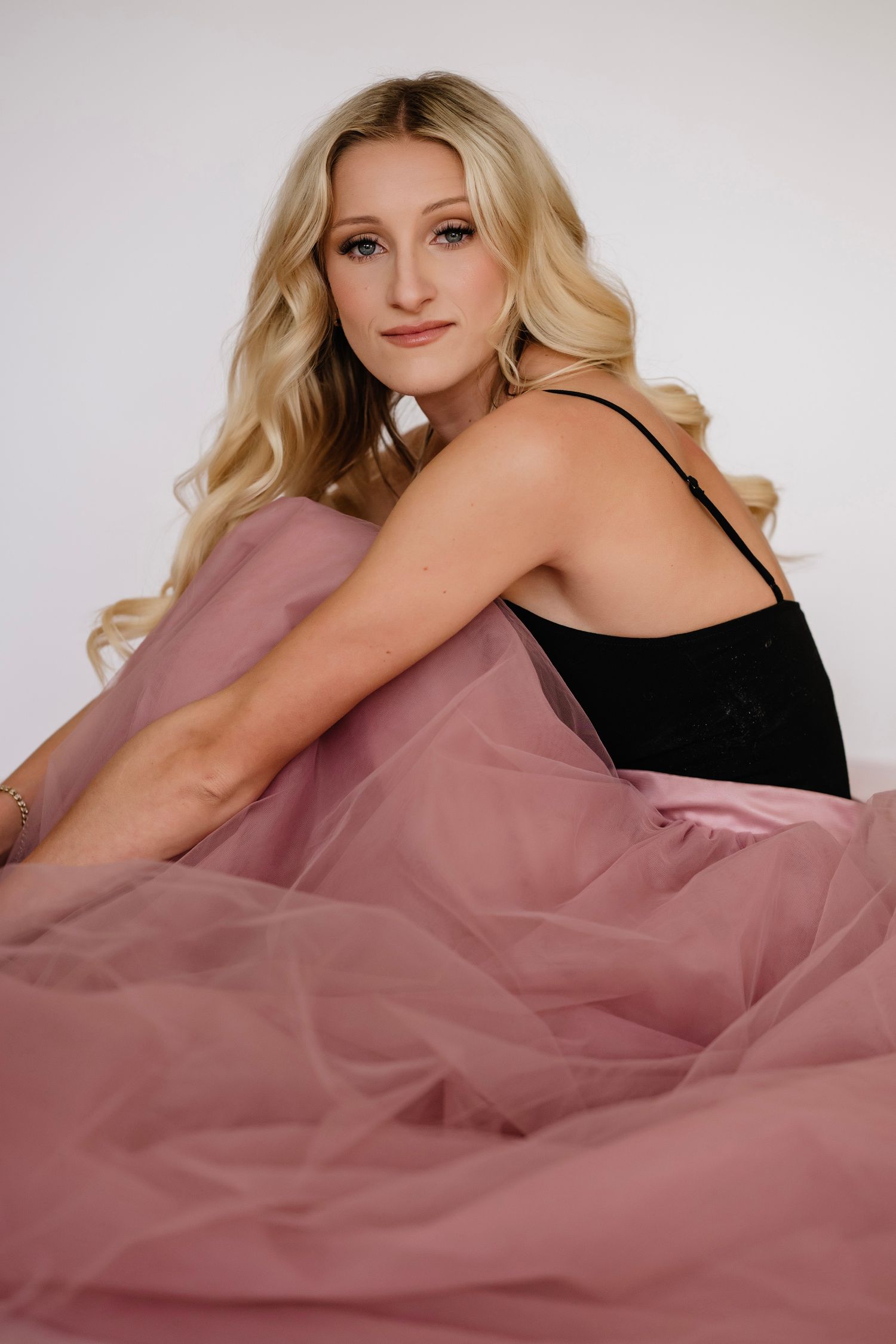 Blonde woman in black bodysuit poses gracefully with flowing pink tulle skirt against white background.