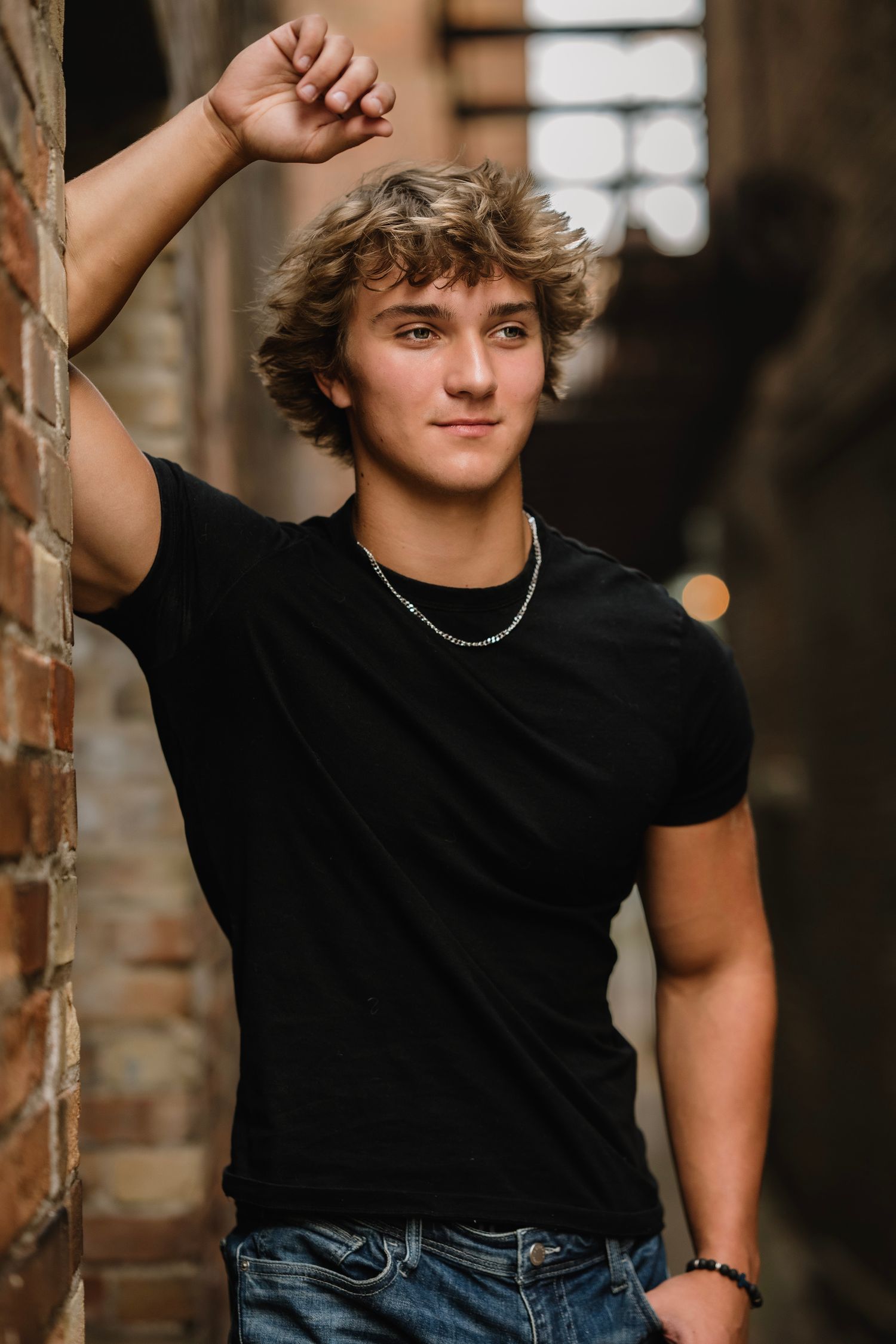 Young person in black t-shirt and jeans poses casually against rustic brick wall in natural lighting.