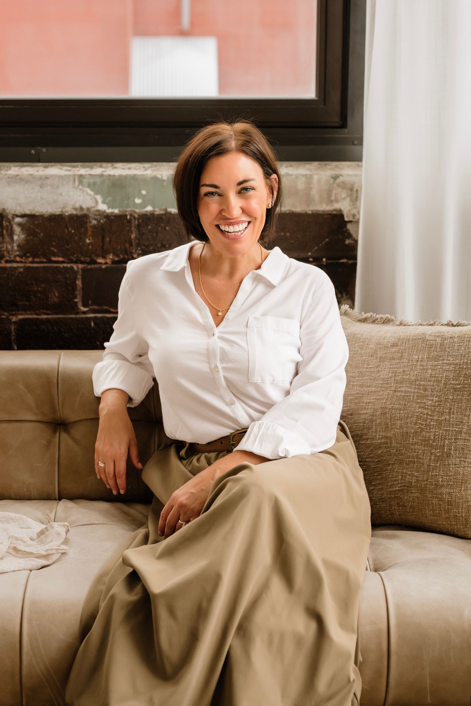 Smiling woman in white shirt and khaki skirt sitting on a beige sofa near a window.