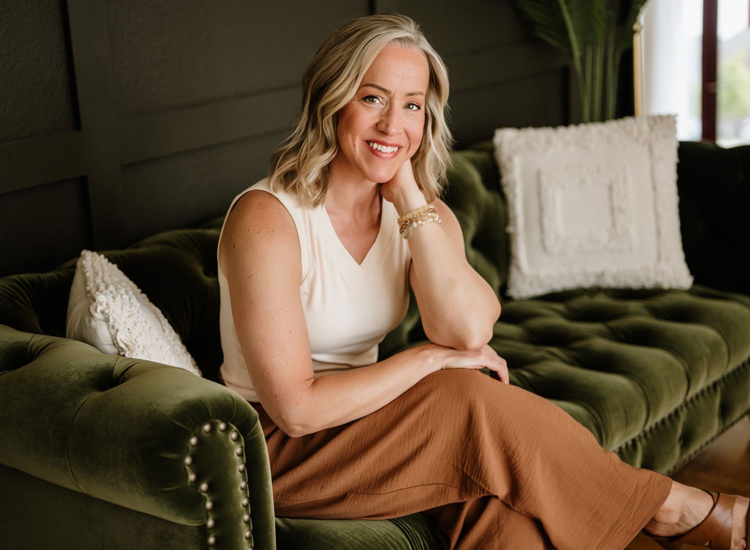 Person lounges in a cozy pose on an olive green velvet couch with natural lighting through window.