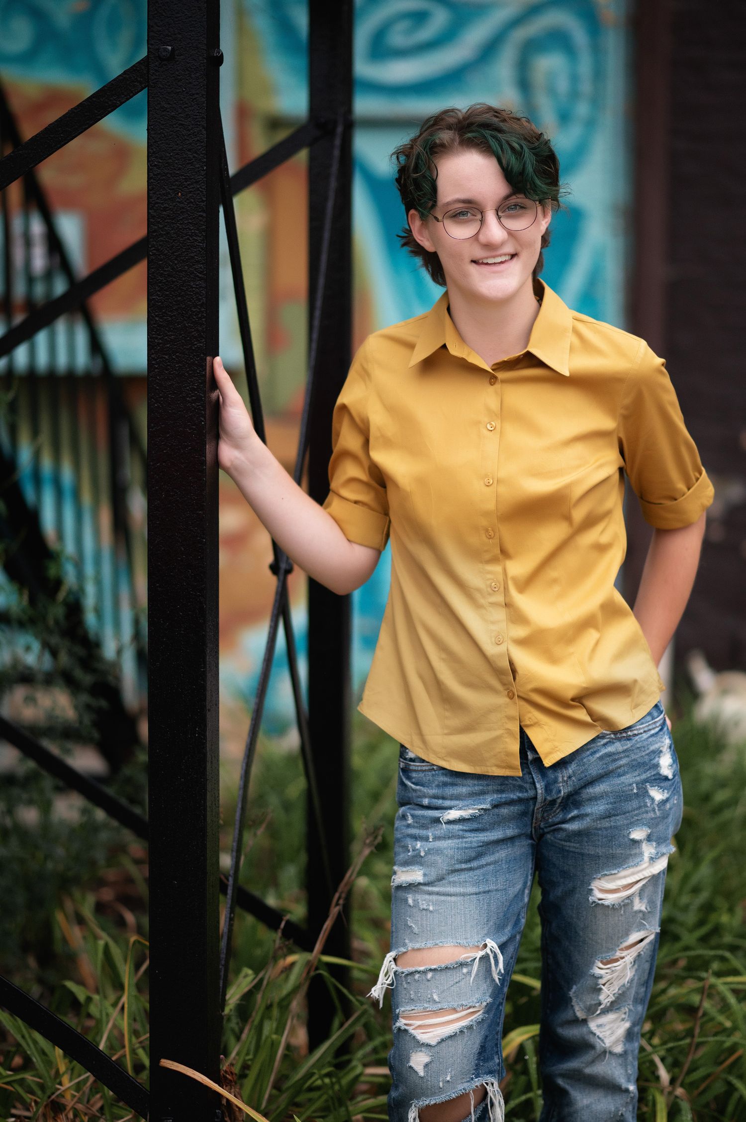 Smiling young person with green hair and glasses wearing a yellow shirt and ripped jeans leaning against a black metal structure.