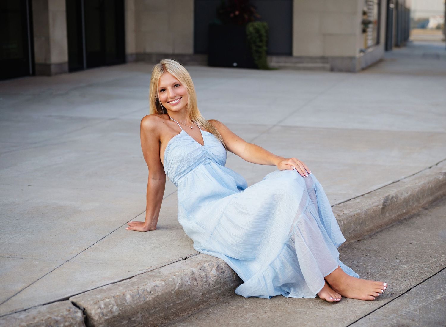 Smiling blonde woman in a light blue maxi dress sitting barefoot on a curb outdoors.