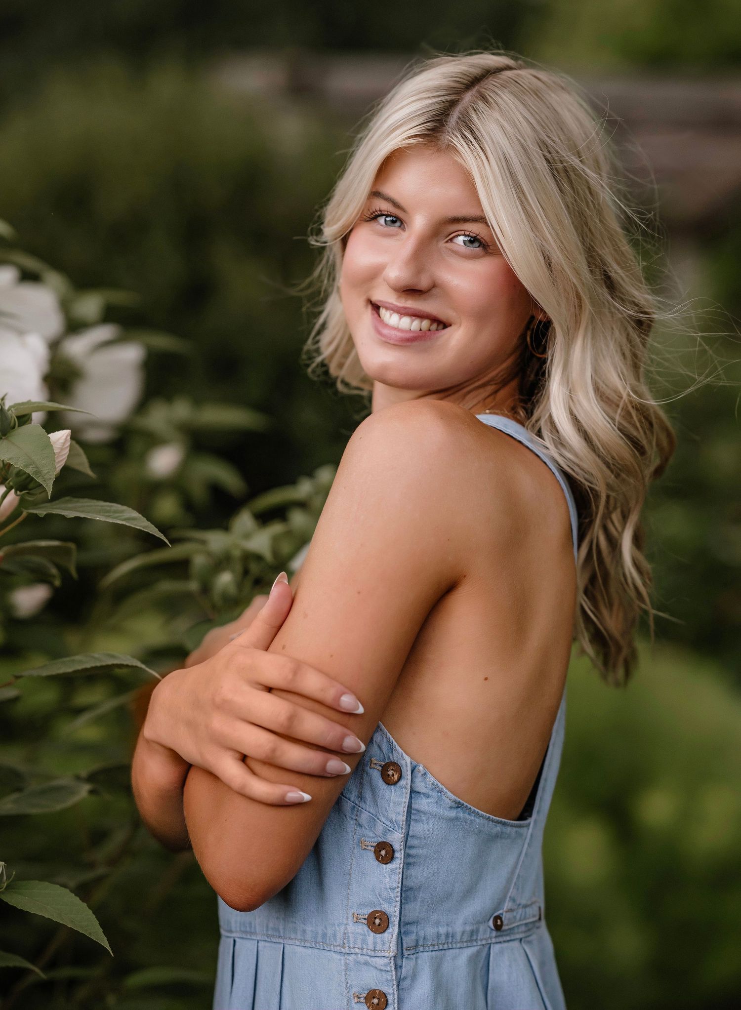 A person wearing a denim overalls dress poses outdoors against a blurred green garden background.