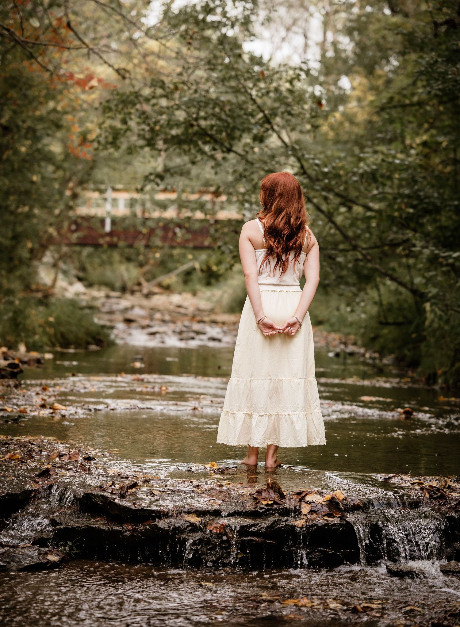 A person in a white dress stands contemplatively at the edge of a rocky stream in an autumn forest setting.