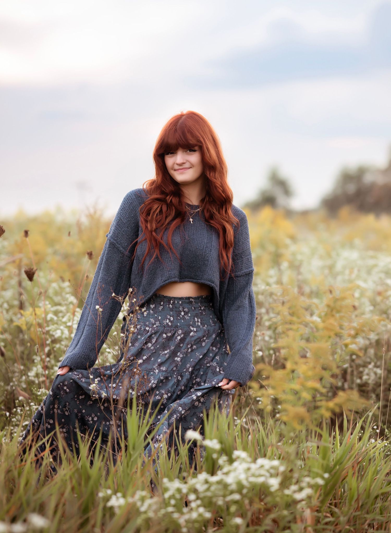 A woman with red hair sits in a field of wildflowers wearing a grey sweater and patterned dress.