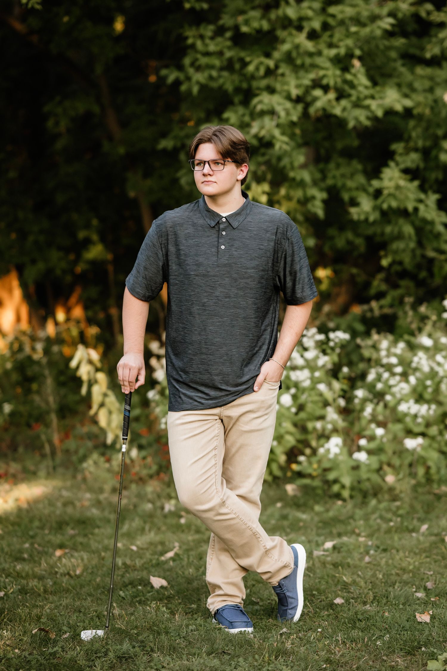 Person in a grey henley shirt and khaki pants walking on grass with trees and white flowers in the background.