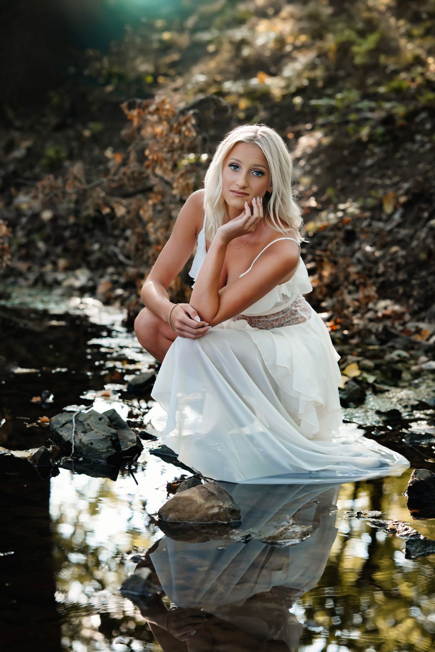 Woman in flowing white dress poses gracefully by reflective pond in autumn woodland setting.