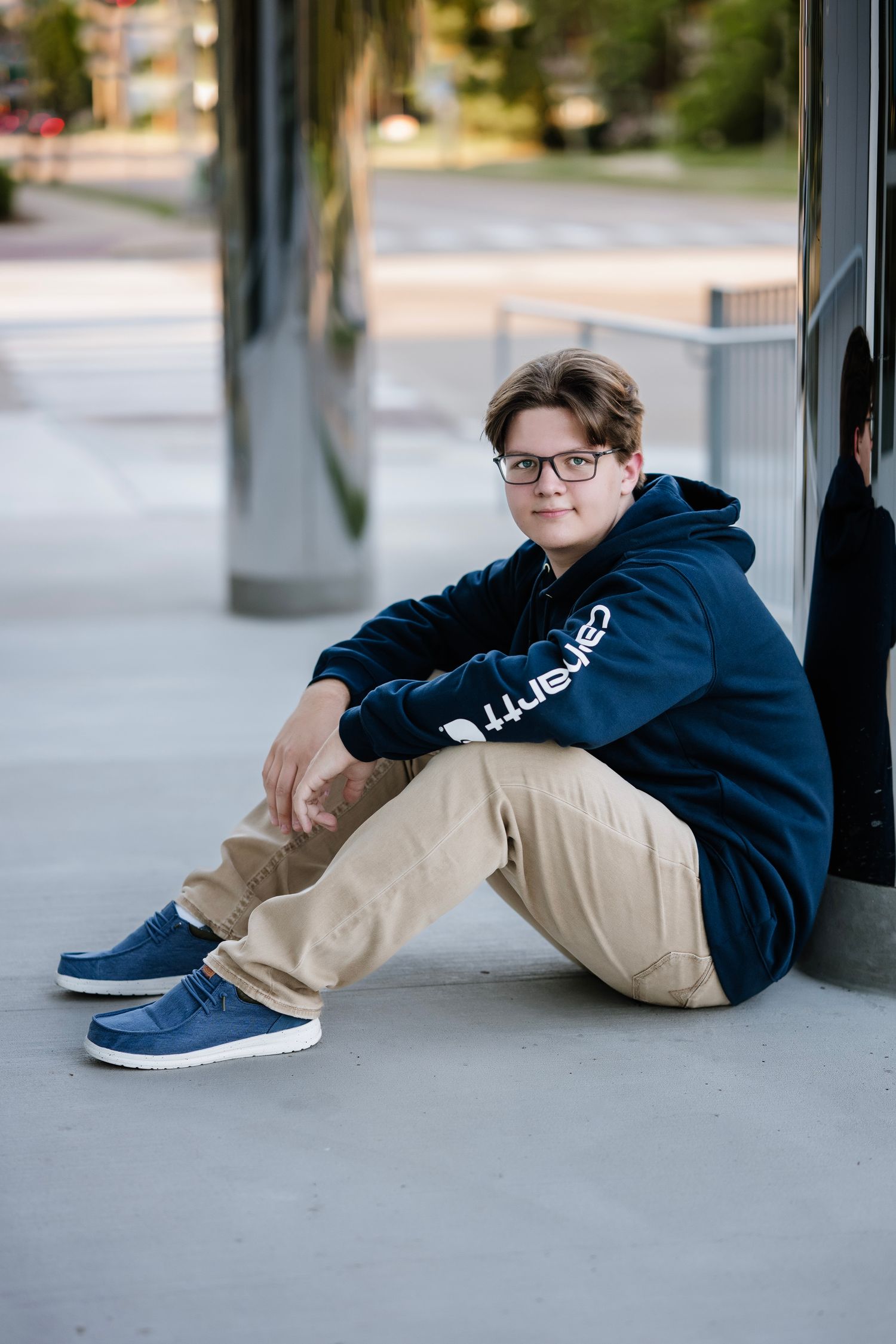 A person in glasses wearing a navy blue hoodie and khaki pants sits casually on a concrete floor with blue sneakers.