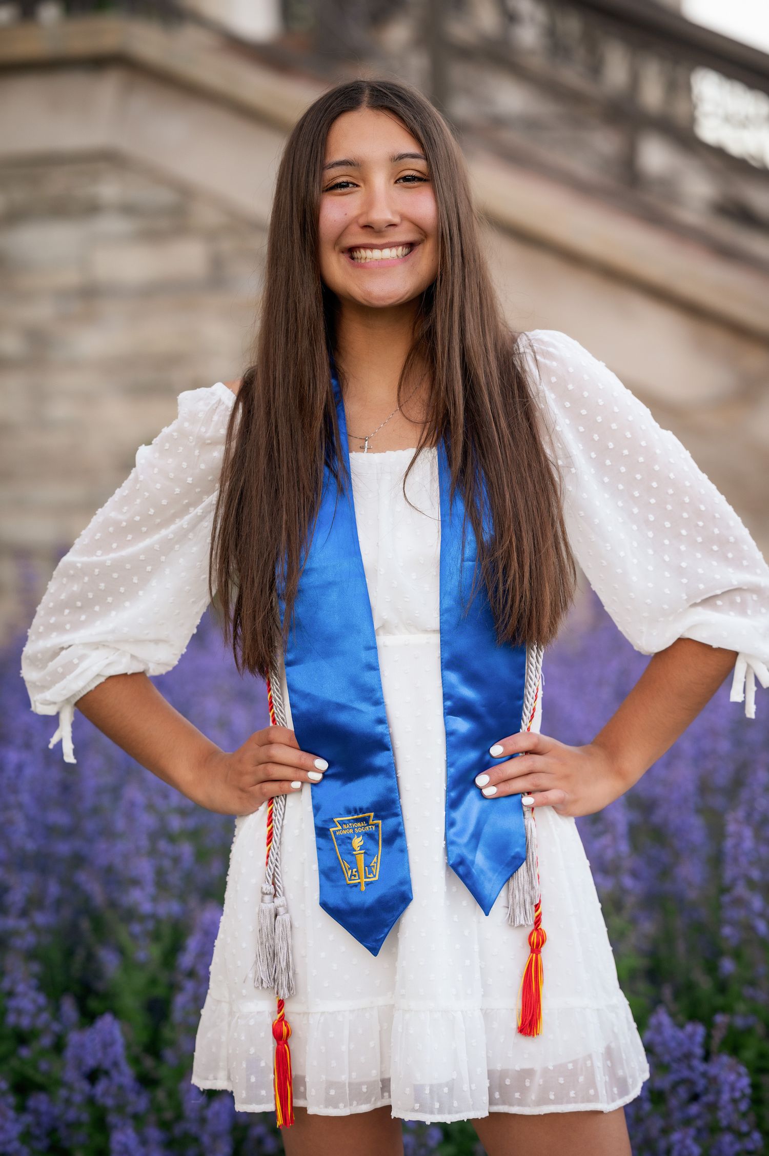 A graduate poses in a white lace dress and blue stole with red tassels against a backdrop of purple flowers.