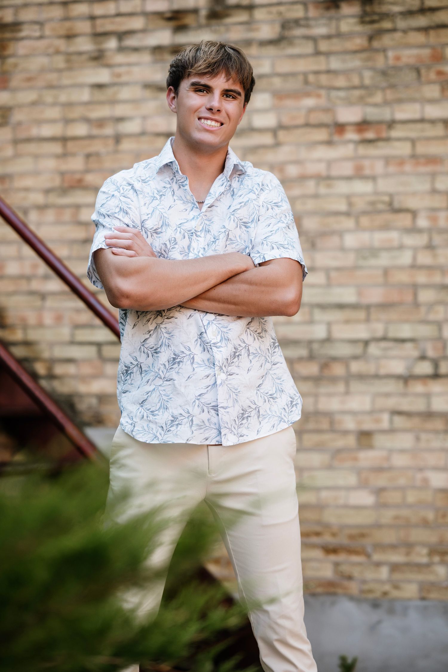 A person wearing a white floral print short-sleeve shirt and khaki pants stands with arms crossed against a brick wall.