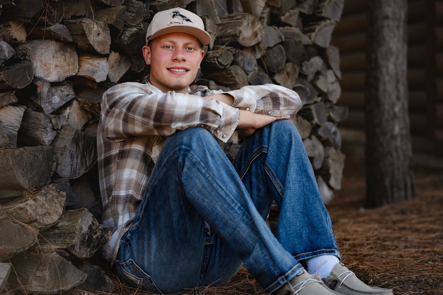 Person in casual wear sits on stone wall in wooded area wearing baseball cap and jeans during outdoor photoshoot.