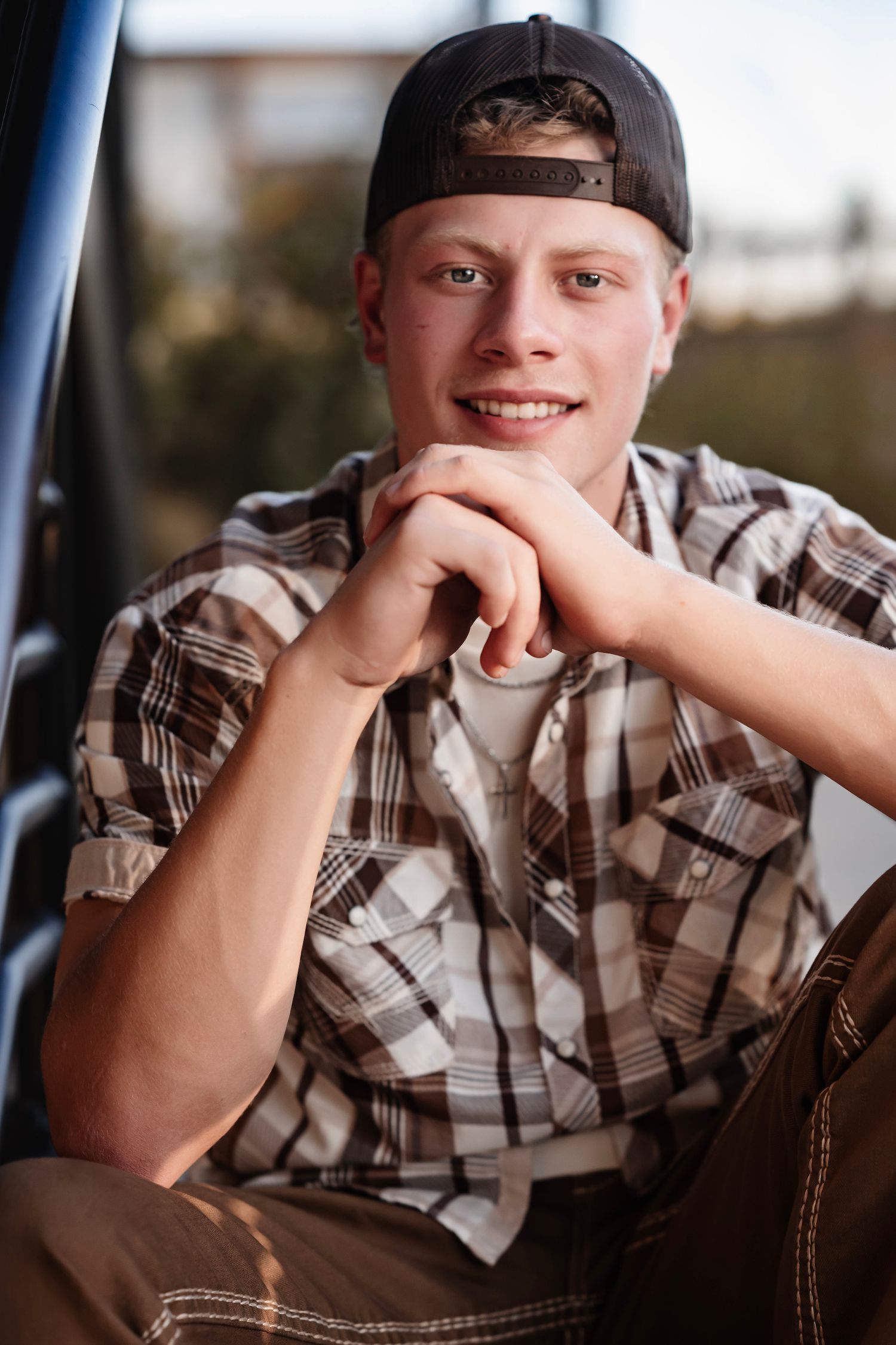 A person in a plaid shirt and black cap poses casually with their hand resting on their chin.