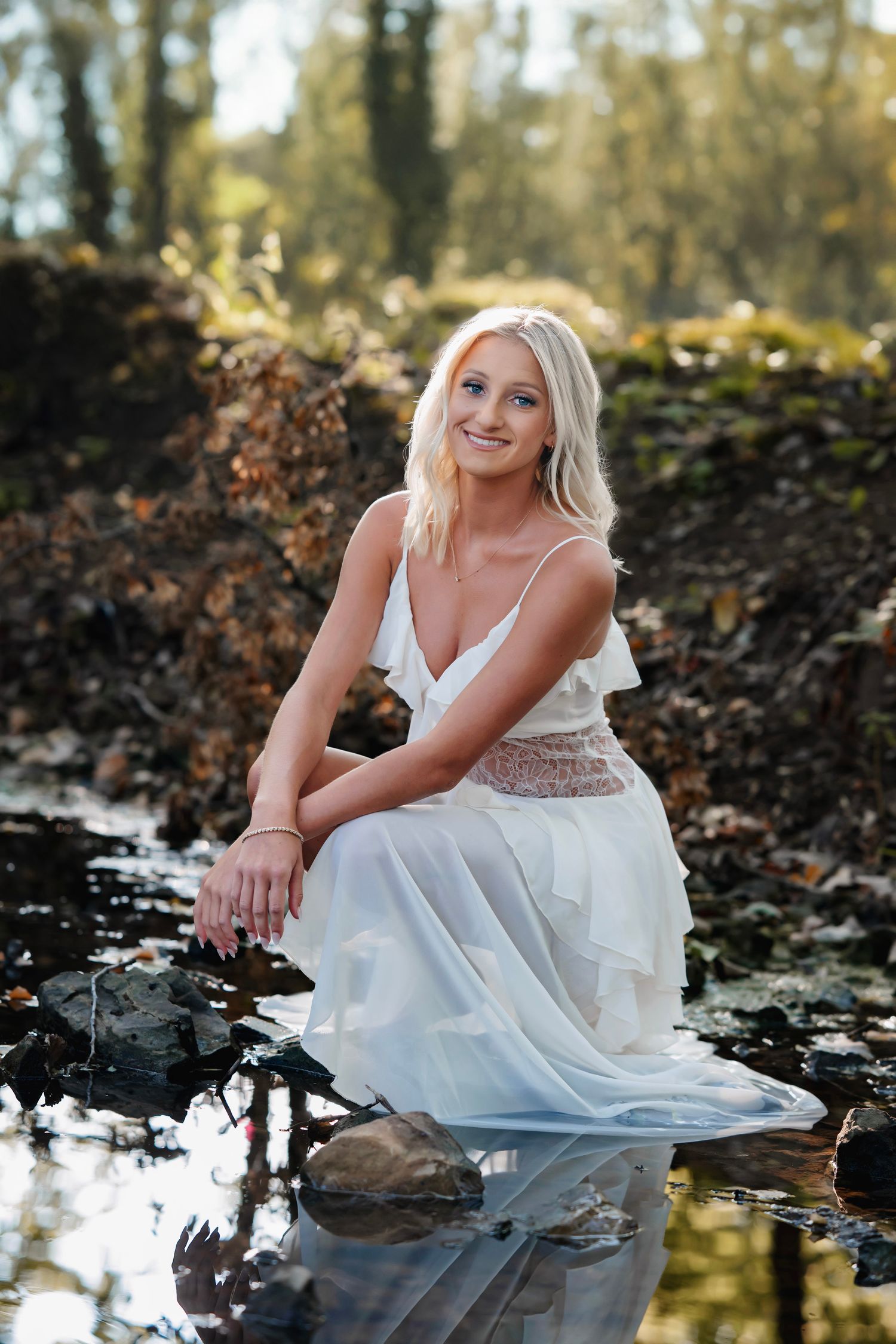 Smiling blonde woman in white dress sitting on rocks by a reflective stream in a sunlit forest setting.