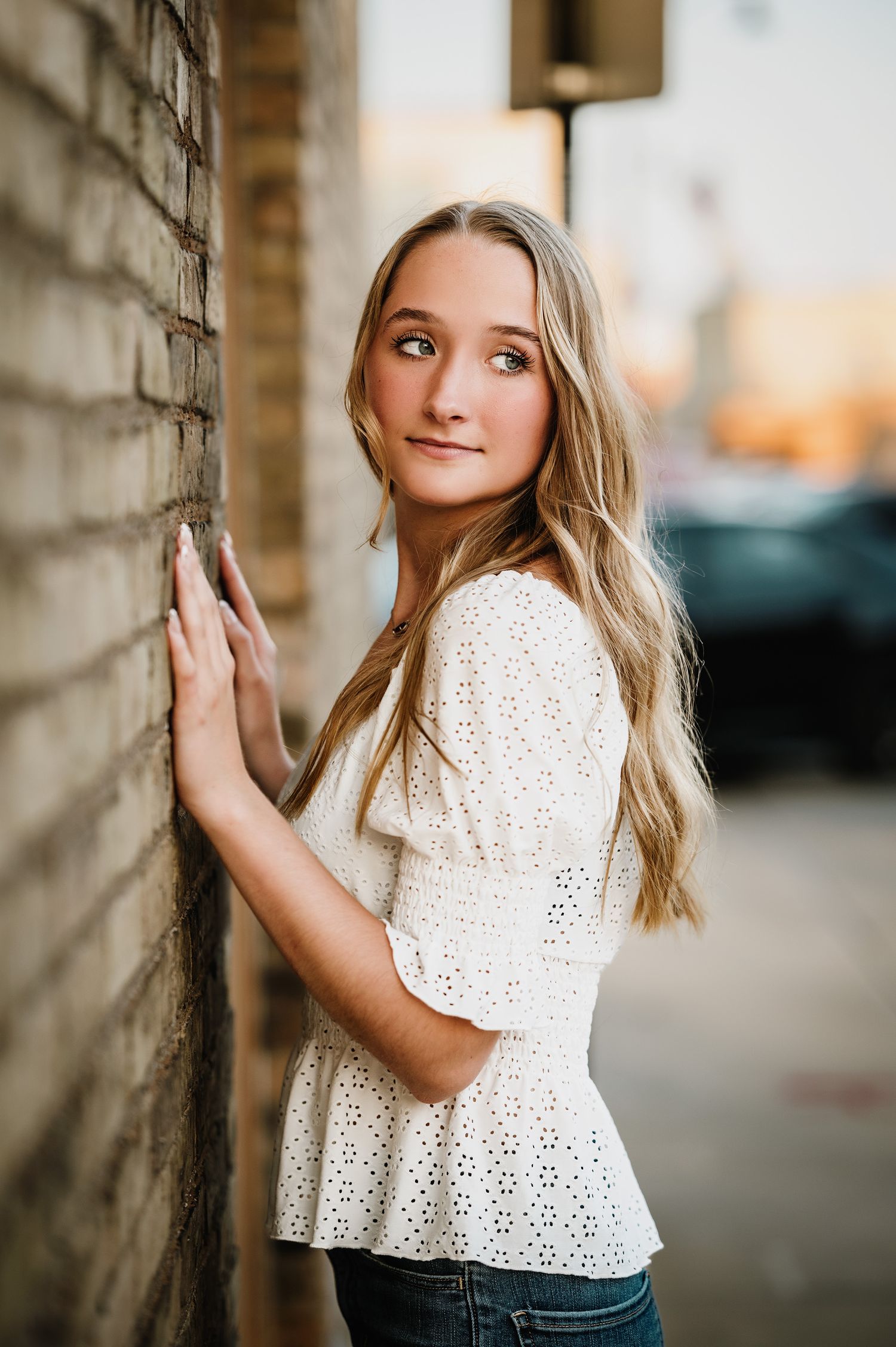 Someone in a white eyelet top poses against a brick wall at sunset.