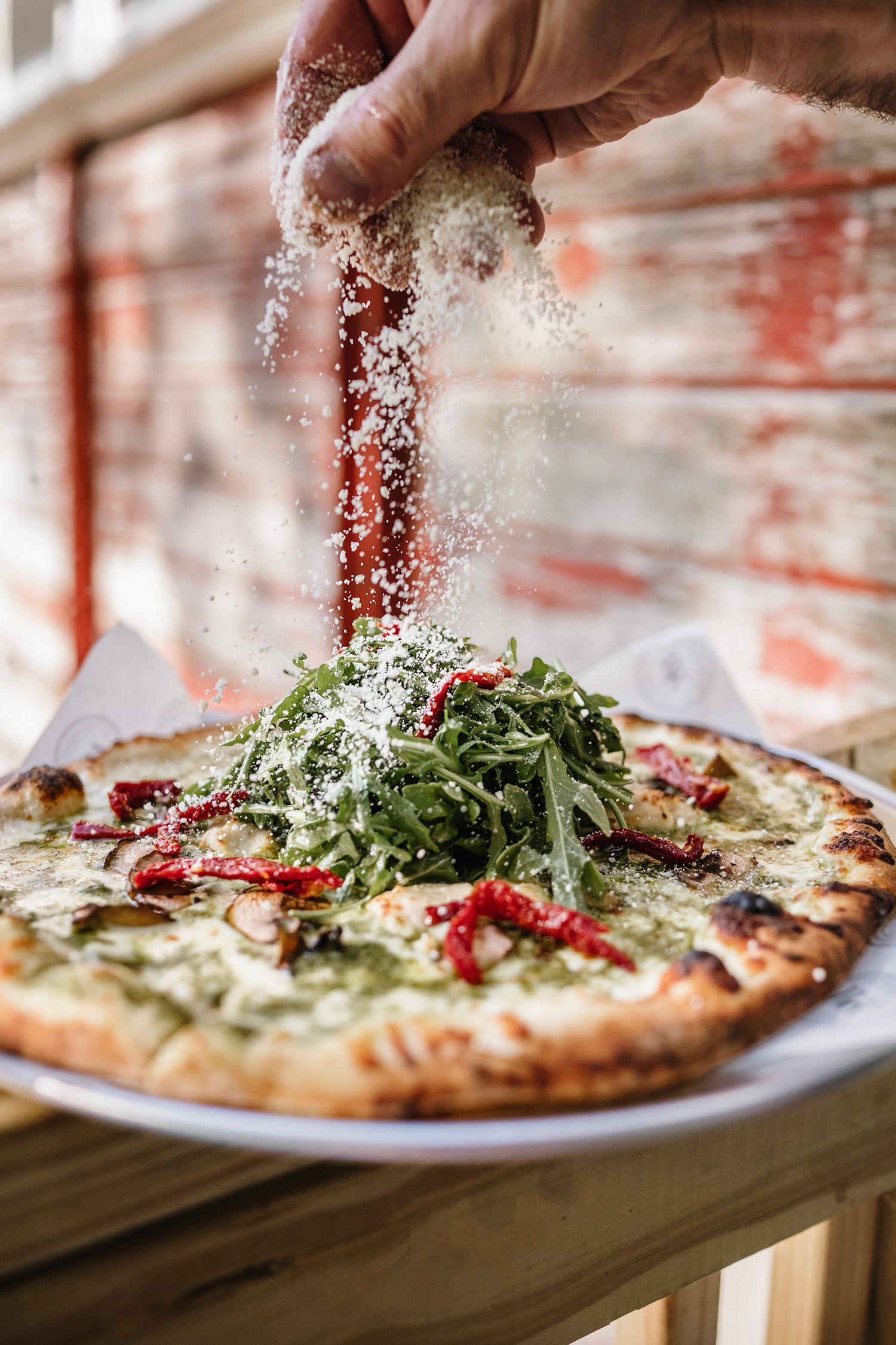 Hand grating fresh parmesan over a wood-fired pizza topped with arugula and sun-dried tomatoes on a white plate.