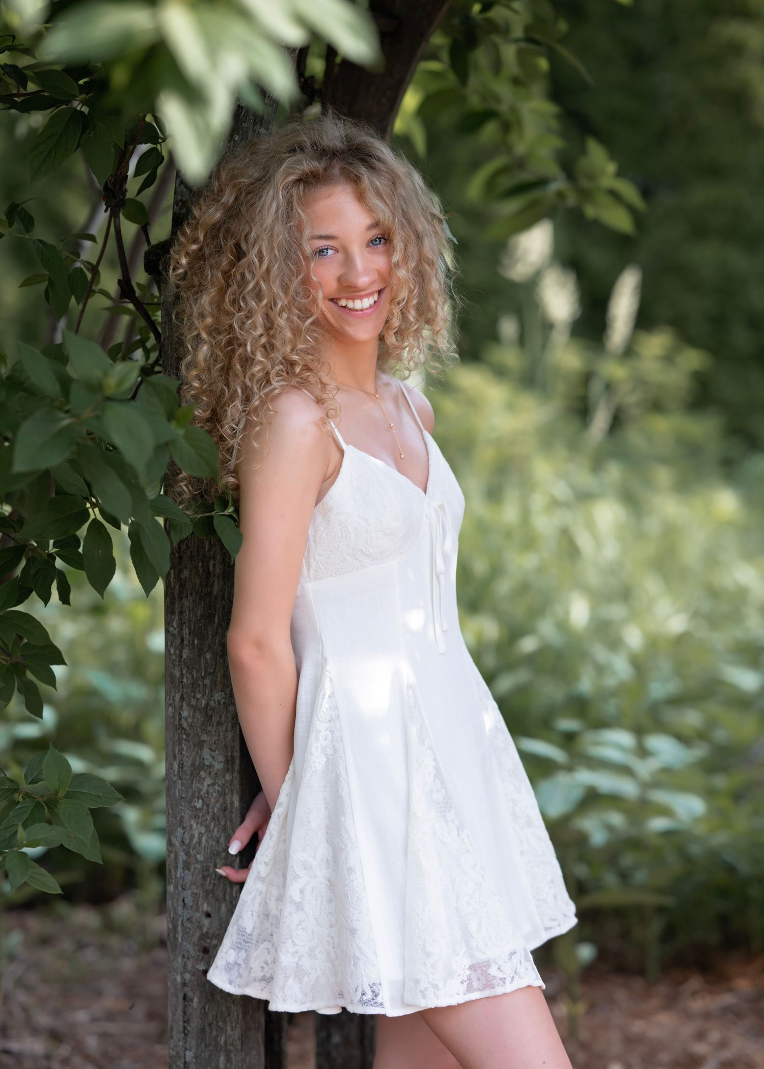 A smiling young woman with curly blonde hair wearing a white lace dress poses outdoors near a tree.