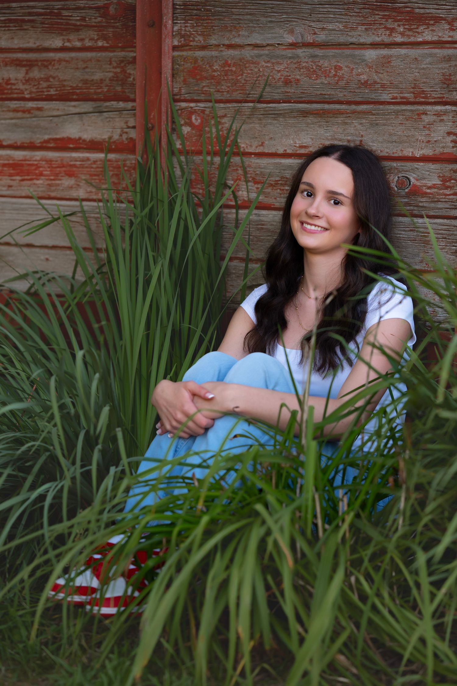 Young woman with dark hair sits smiling among tall green grass against a rustic red barn wall.