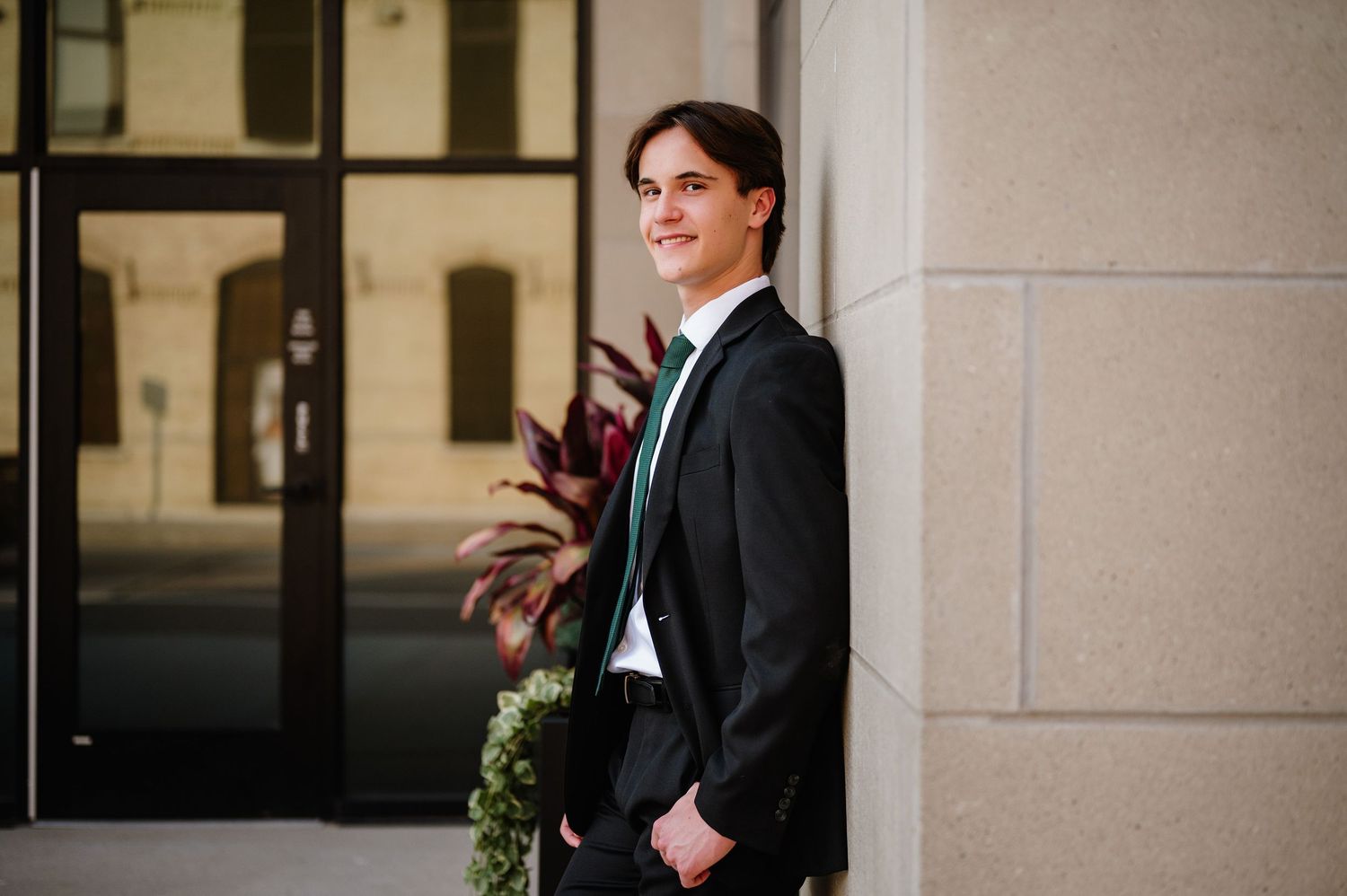 Young man in black suit and green tie leaning against a column outside a building.