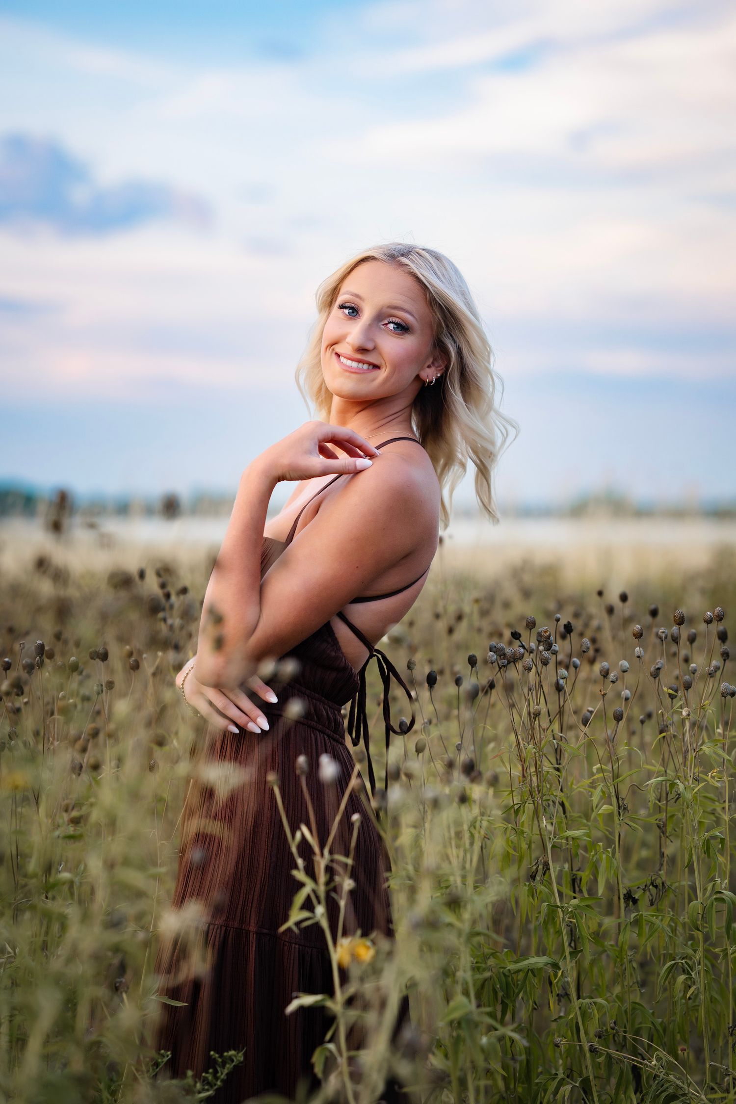 A woman in a brown dress poses joyfully in a field of dried wildflowers during golden hour.