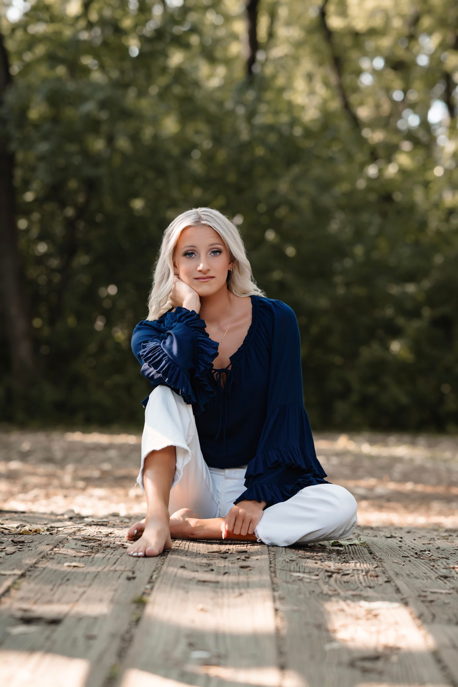 A person in a navy blue top and white pants poses casually on a wooden deck in dappled sunlight.