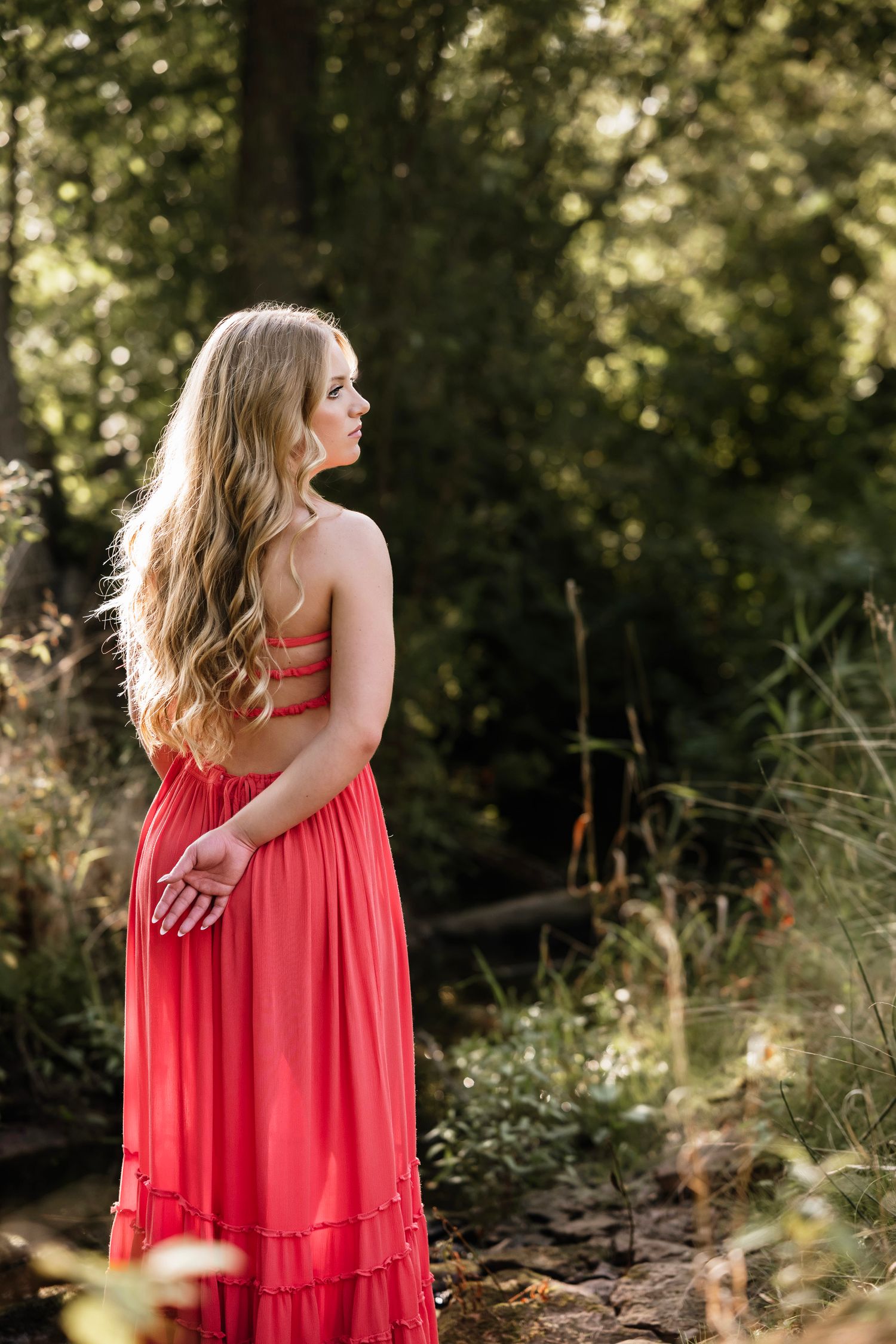Young woman in coral maxi dress standing in lush green forest, long blonde hair, looking away gracefully.