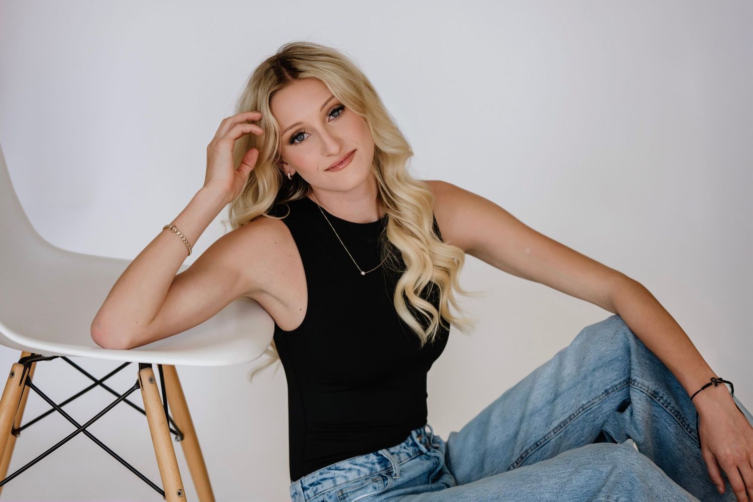 High school senior in black tank top and jeans poses casually on white modern chair against white background.