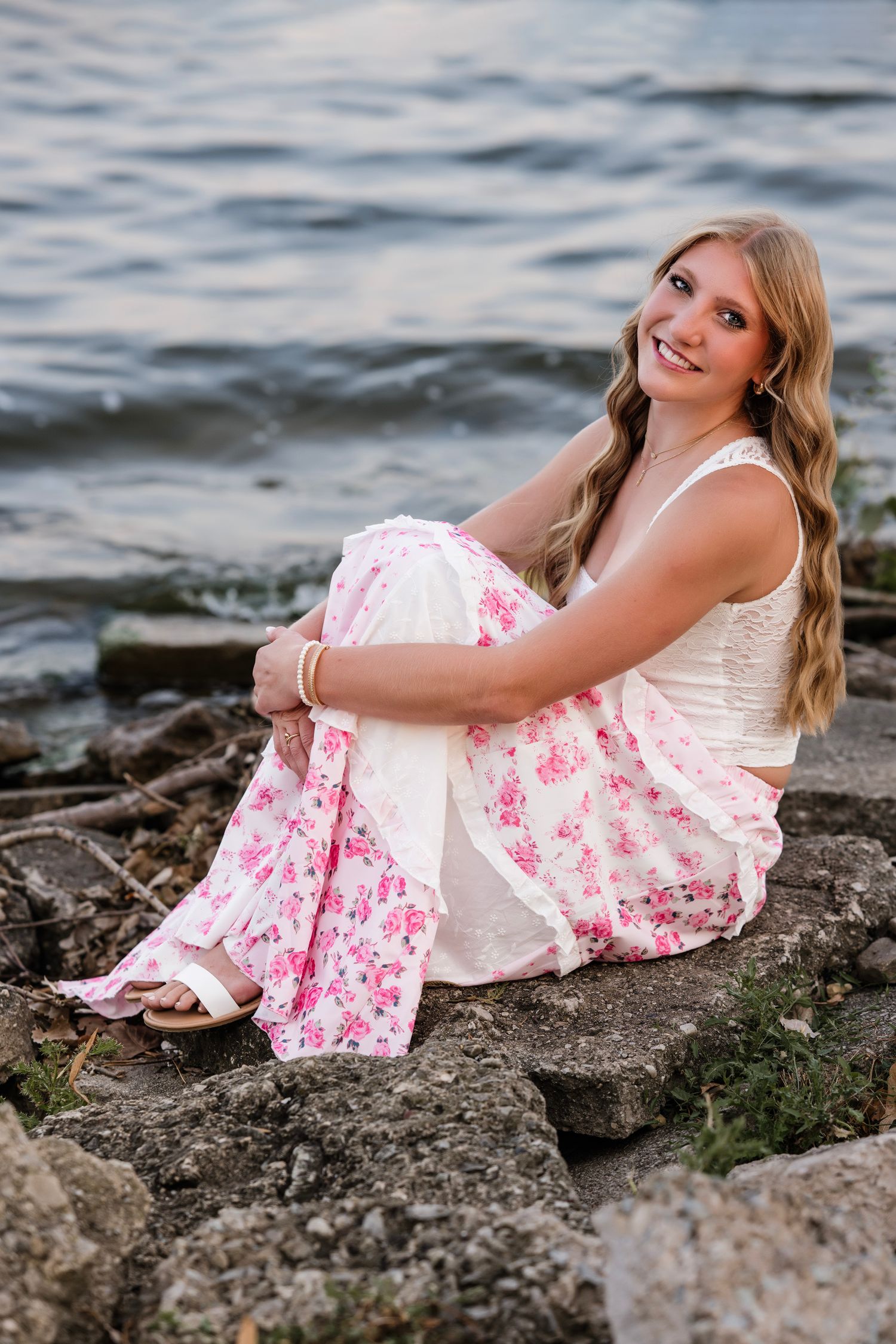 Smiling young woman in floral skirt and white top sitting on rocky shoreline by water.