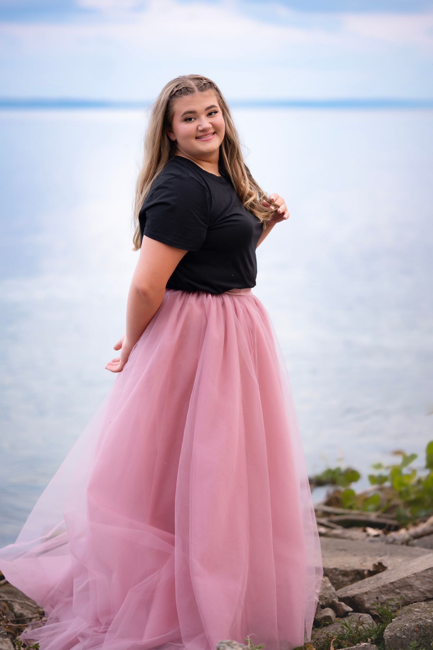 Young woman in black top and pink tulle skirt posing by the water on a rocky shoreline.