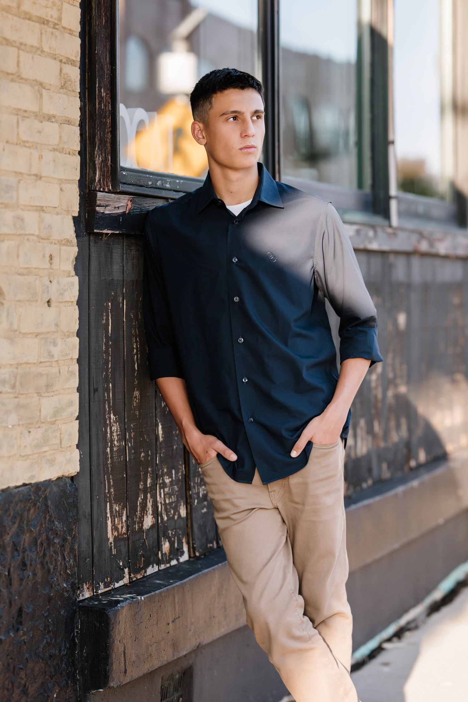 A person in a navy polo shirt and khaki pants leans against a brick wall with windows in a casual pose.