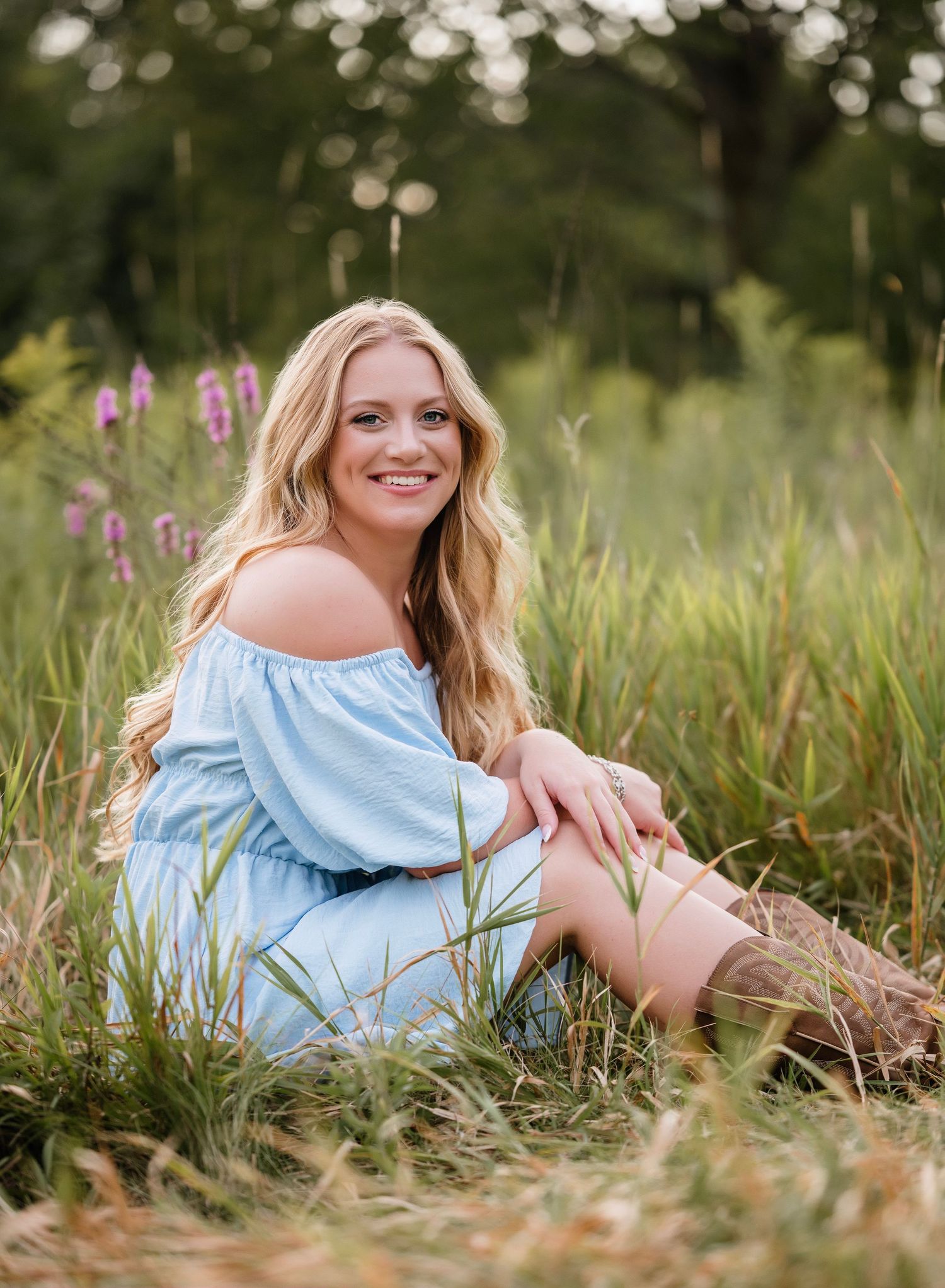 A person in a light blue off-shoulder dress sits in a meadow with purple wildflowers and bokeh-lit trees in background.