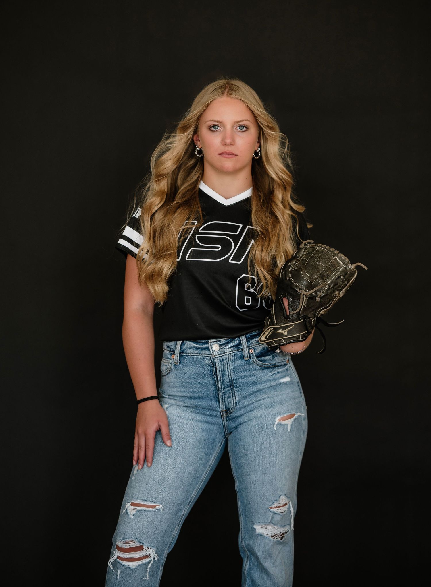 A person in a black athletic jersey and distressed blue jeans poses against a dark background in a casual fashion style.