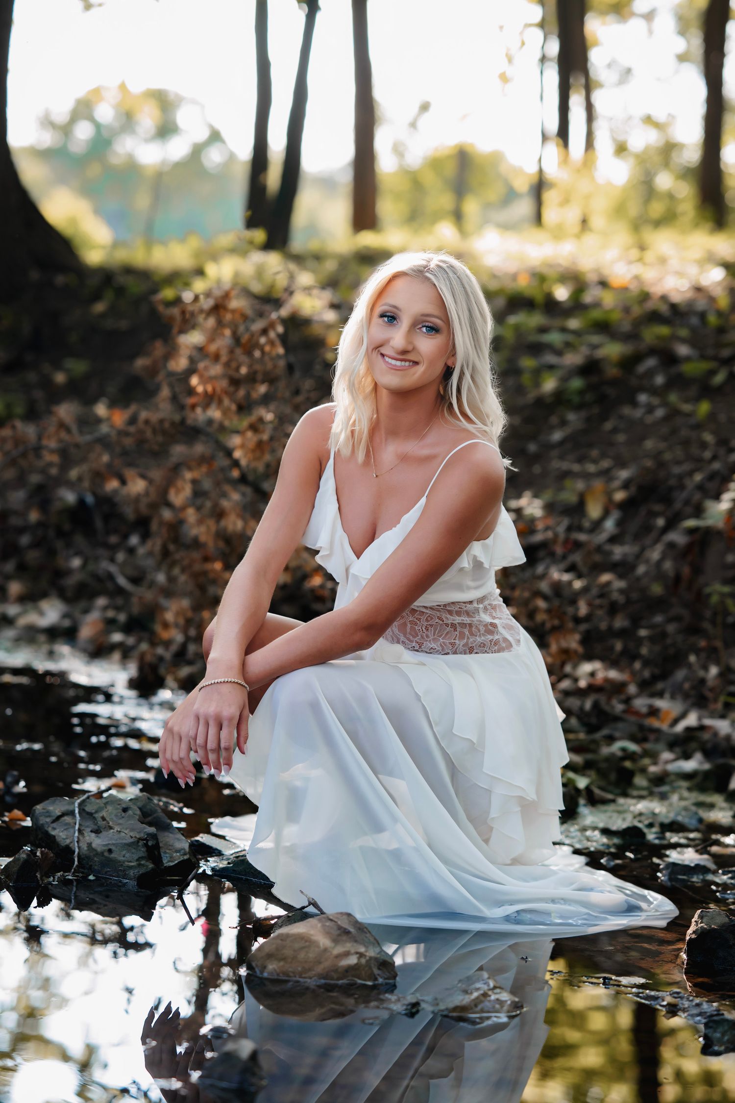 A person in a white dress sits gracefully by a reflective pond in a wooded outdoor setting during golden hour.