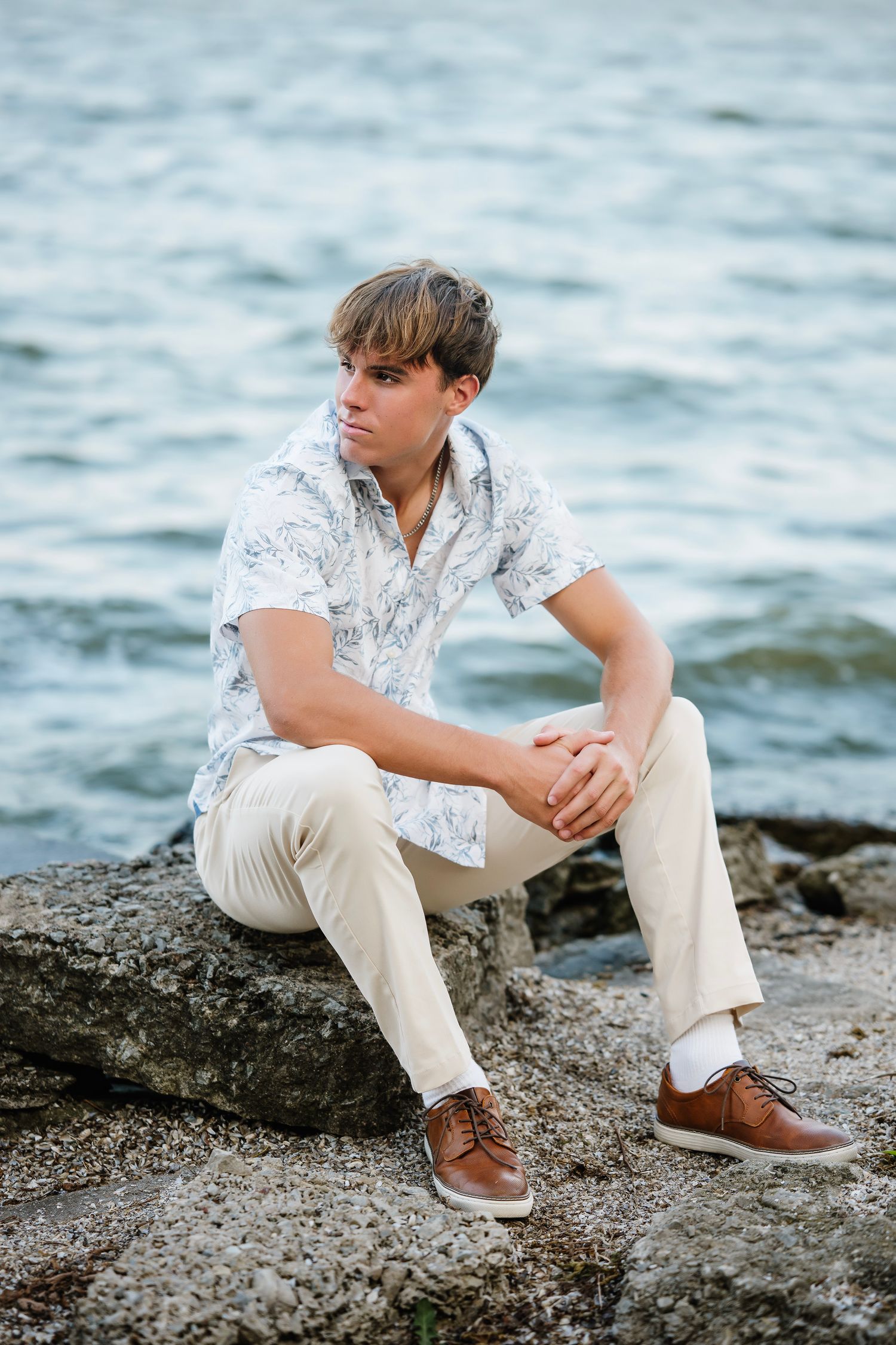 A person in a white patterned shirt and khaki pants sits thoughtfully on rocky shoreline by the water.