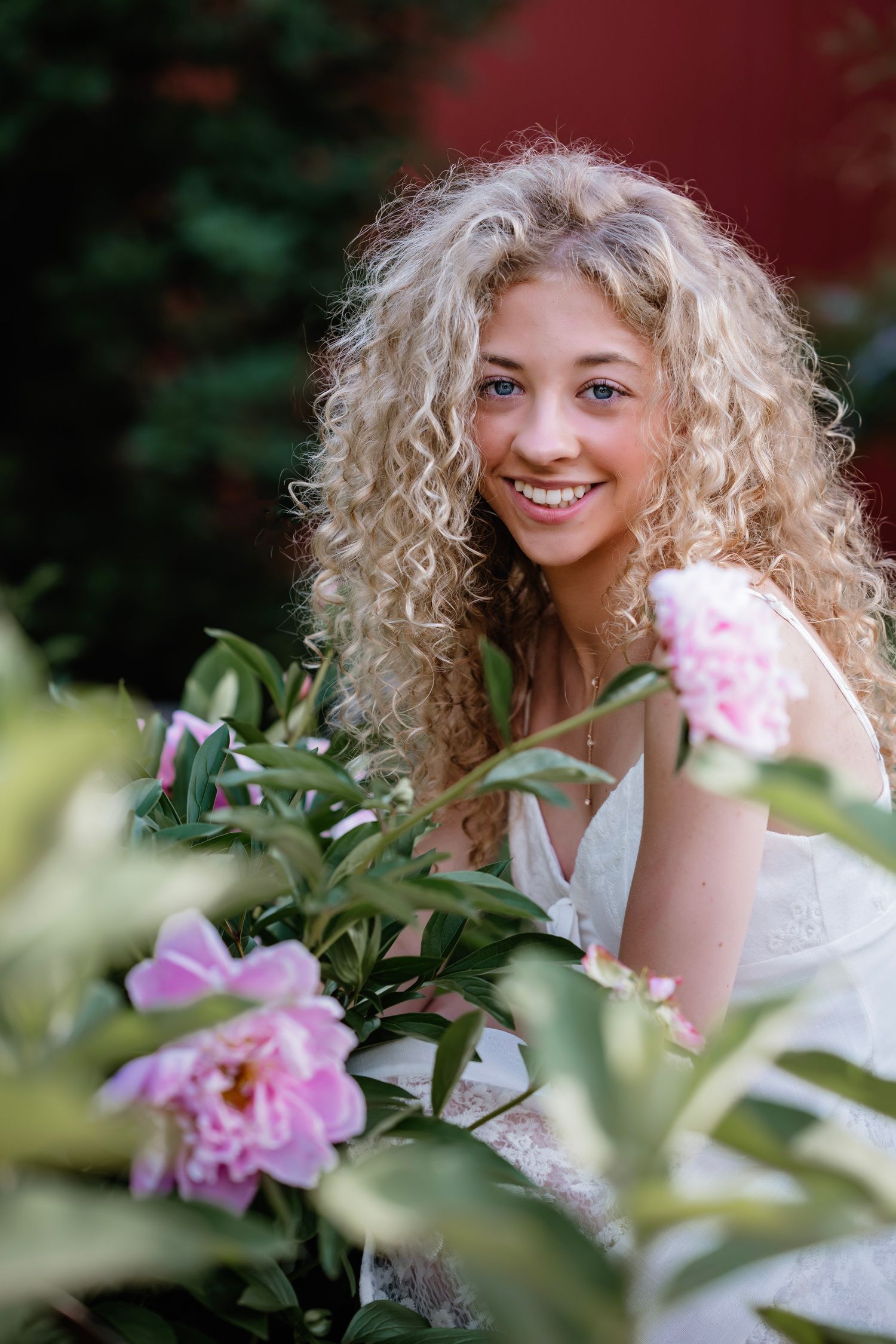 Smiling young woman with curly blonde hair posing among pink peony flowers in a garden wearing a white dress.