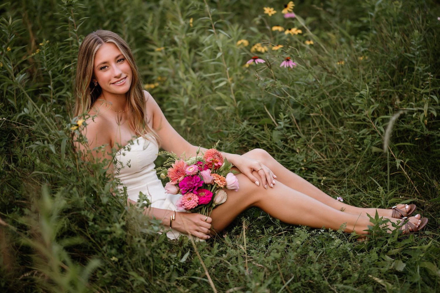 Young woman in white dress sitting in lush green field holding vibrant pink and orange flower bouquet, smiling outdoors.